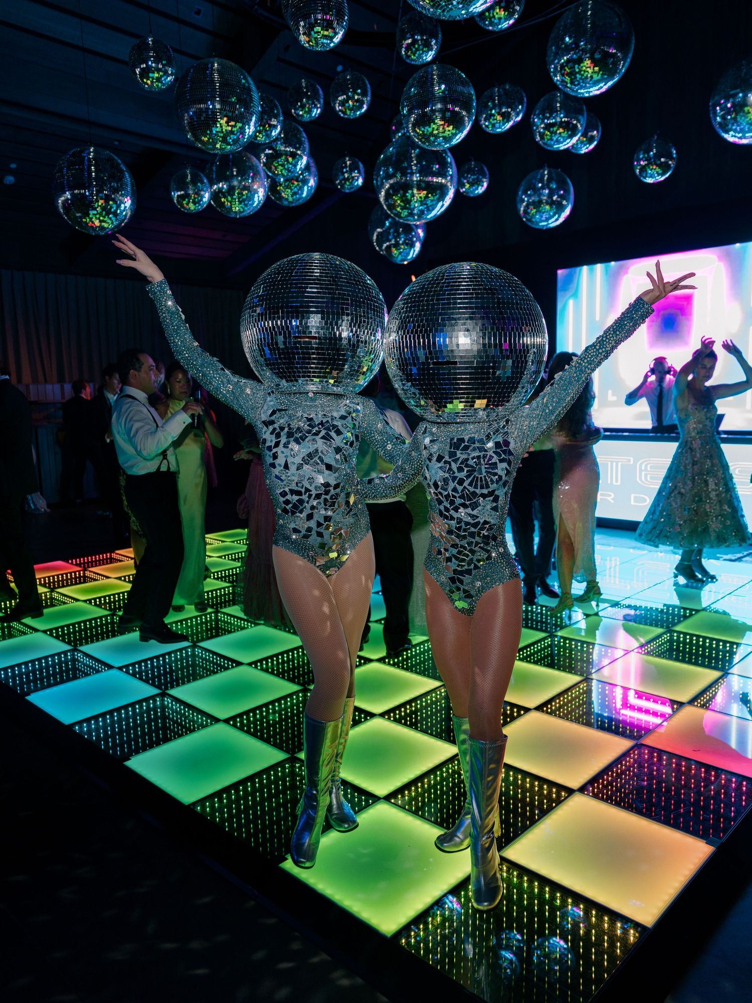Two disco ball-headed performers in silver sequin costumes pose on a glowing LED dance floor.