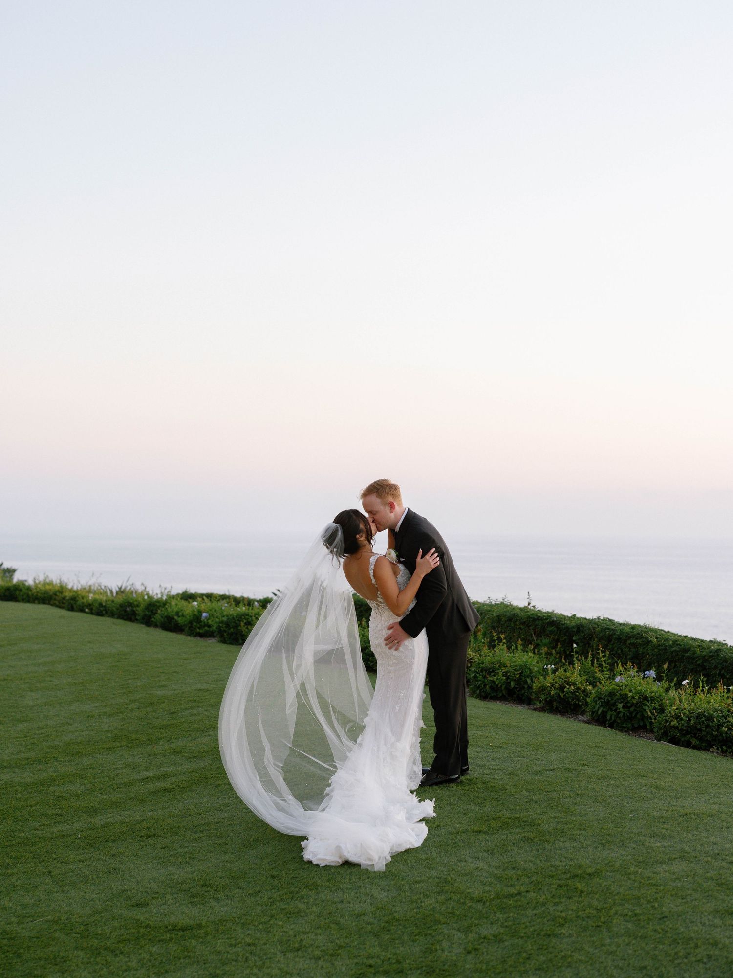 Bride and groom share a kiss at sunset on a green lawn overlooking the ocean, veil flowing in the breeze.