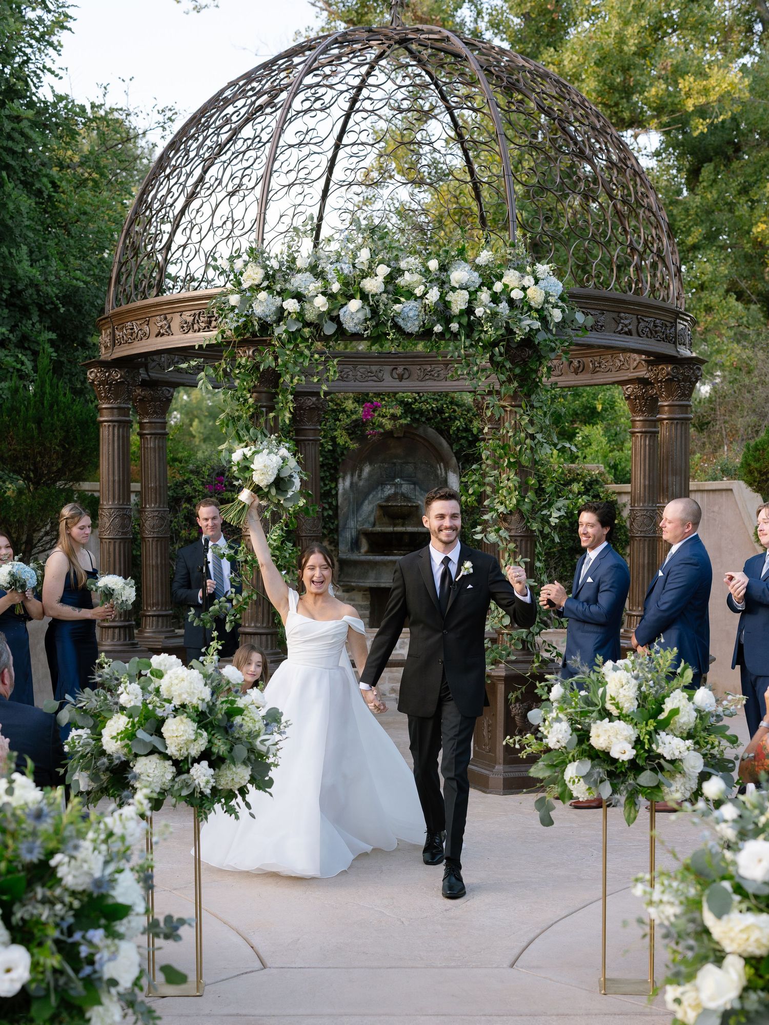 Joyful bride and groom recessional under ornate iron gazebo adorned with white flowers and greenery.