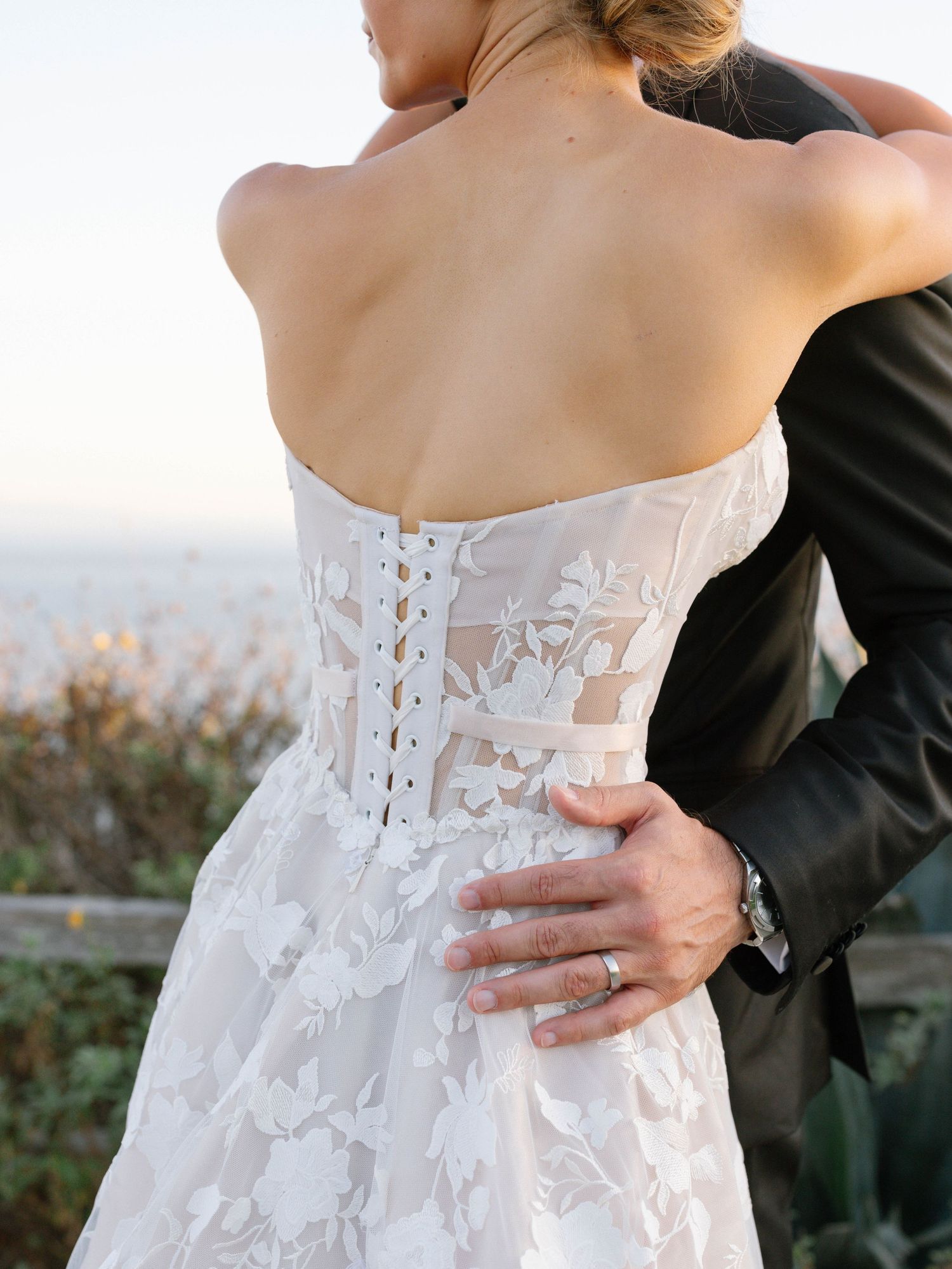 Detail shot of bride's intricate lace corset wedding gown back with button closure, groom embracing her.