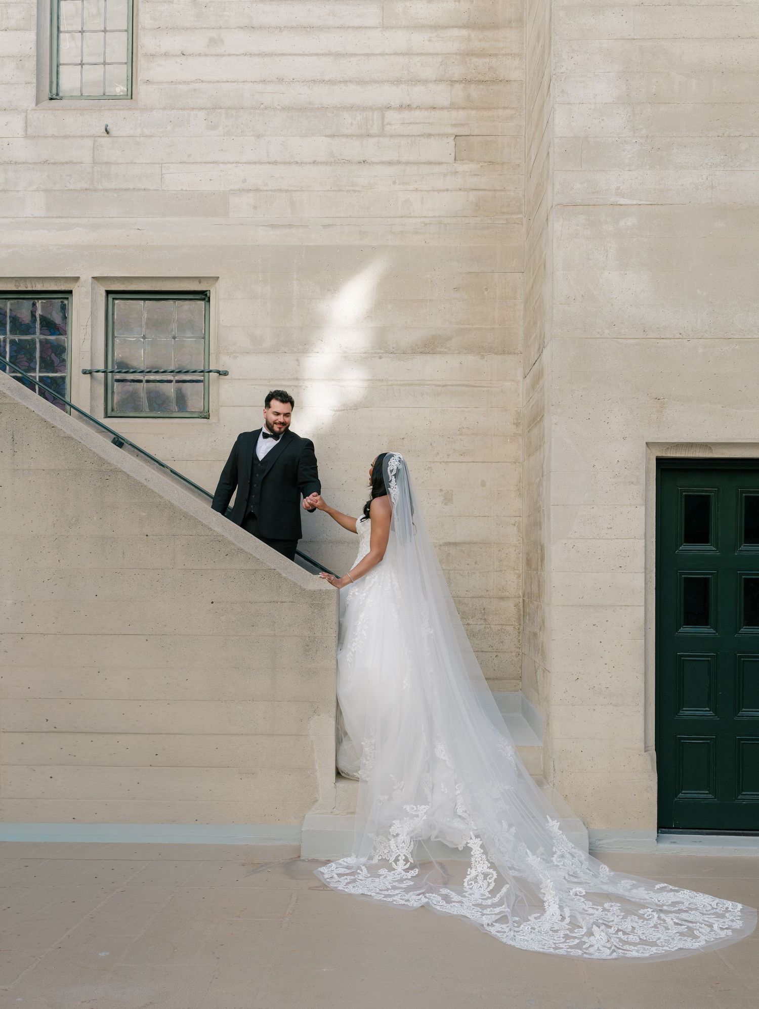 Bride in lace gown with cathedral veil holds grooms hand on stone staircase at wedding.
