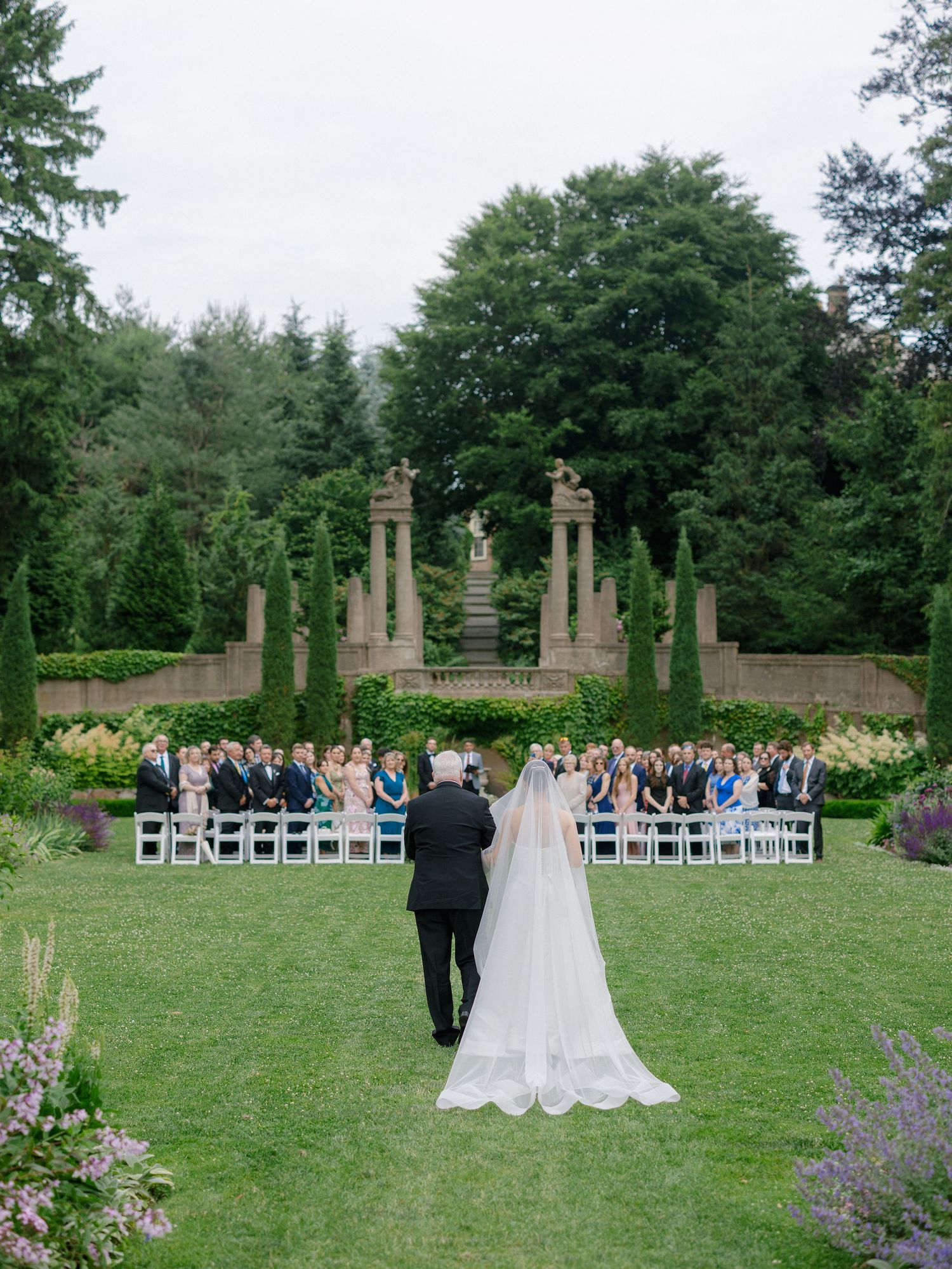 Bride and escort walking toward outdoor wedding ceremony in lush formal garden setting.