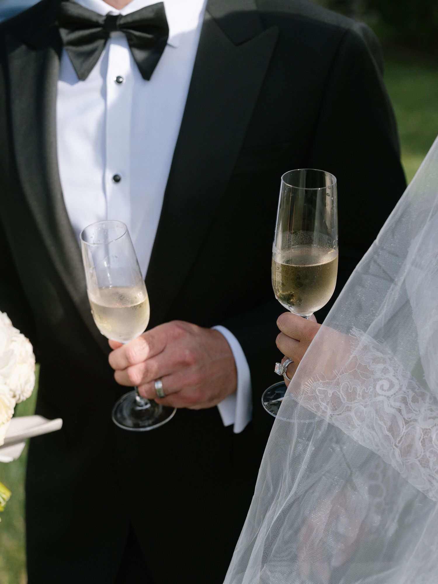 Bride and groom in black tuxedo and white veil toasting with champagne flutes at outdoor wedding.