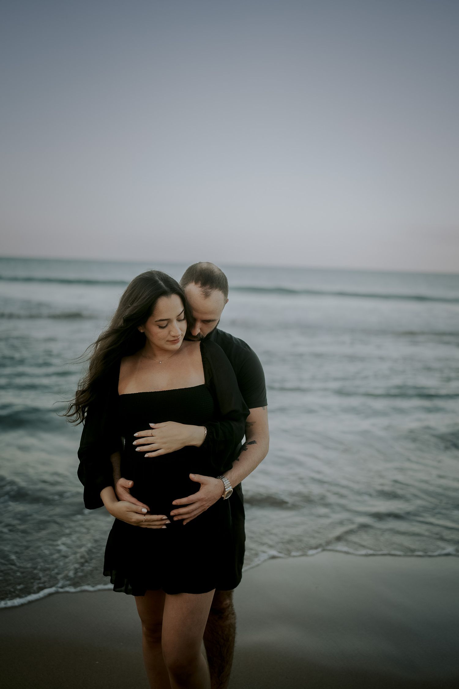 Couple in black outfits embracing on beach at dusk, man tenderly holding pregnant woman's belly near ocean waves.