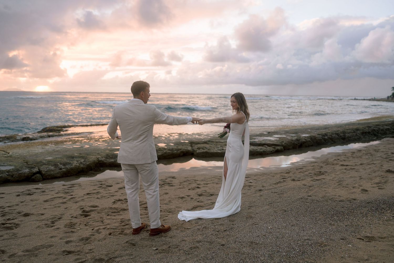 Bride and groom hold hands on sandy beach at sunset during romantic coastal wedding celebration.