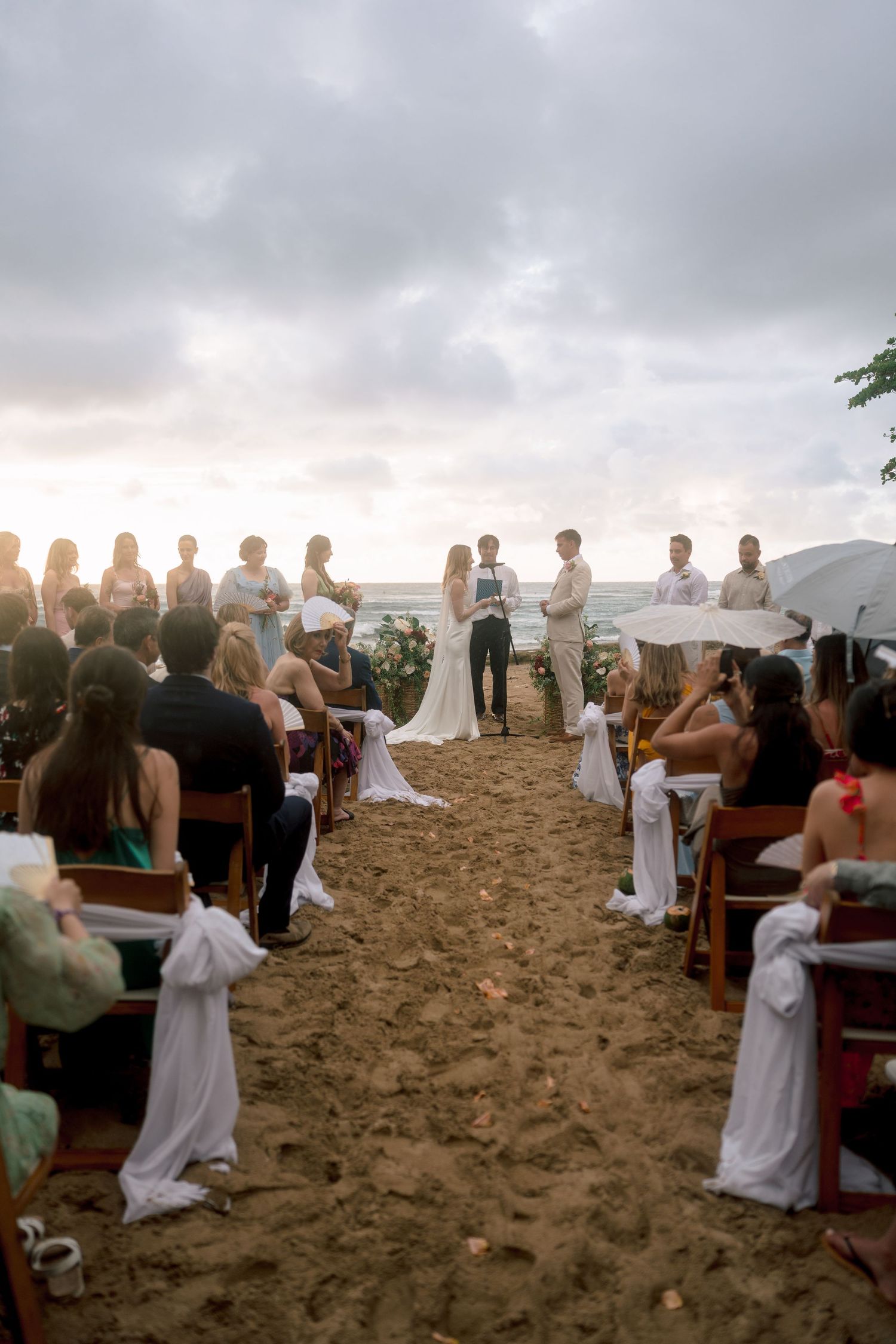 Romantic beach wedding ceremony at sunset with guests seated on wooden chairs along a sandy aisle.