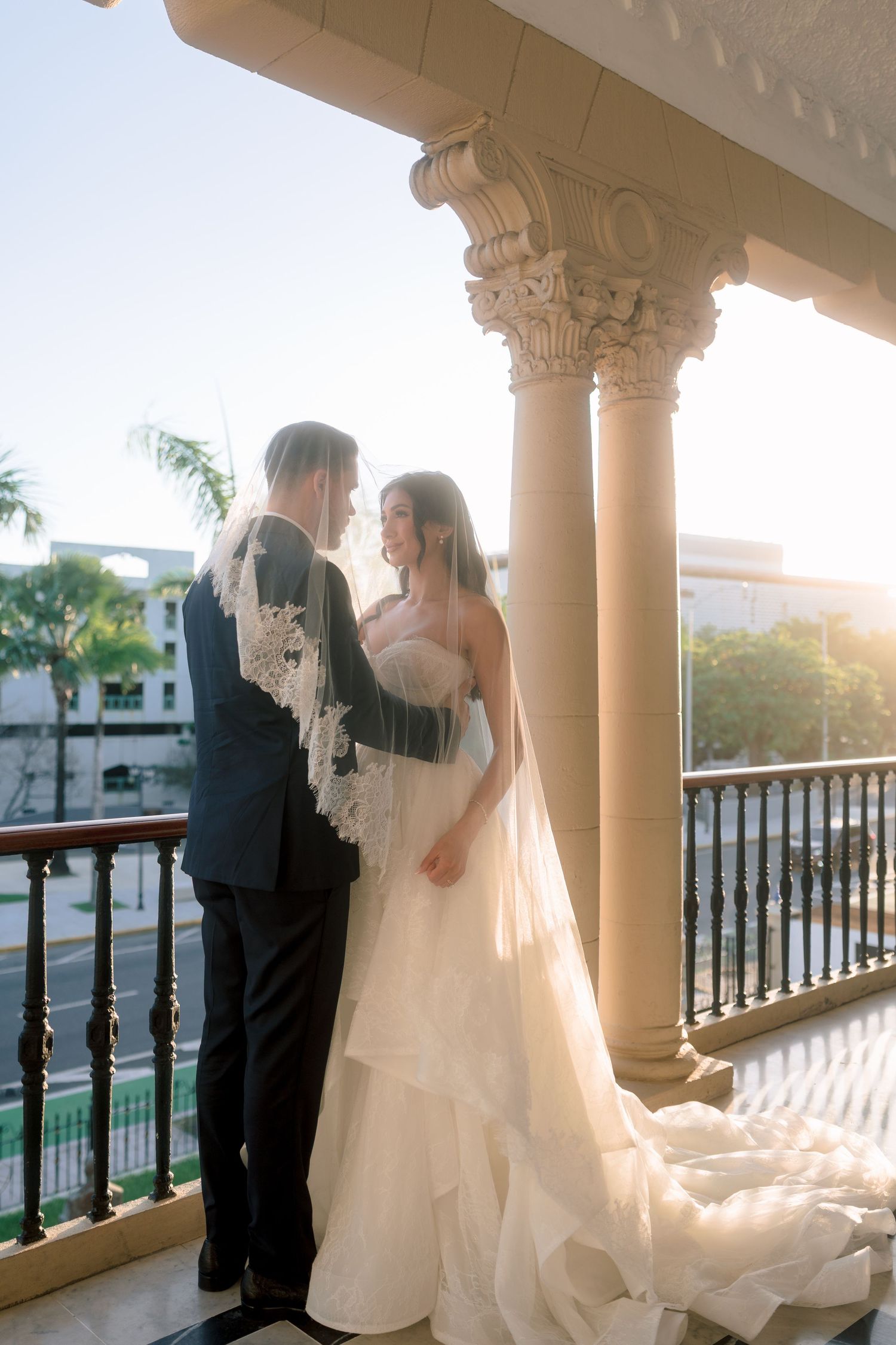 Bride in strapless layered gown and groom embrace on ornate balcony with Corinthian columns at golden hour.