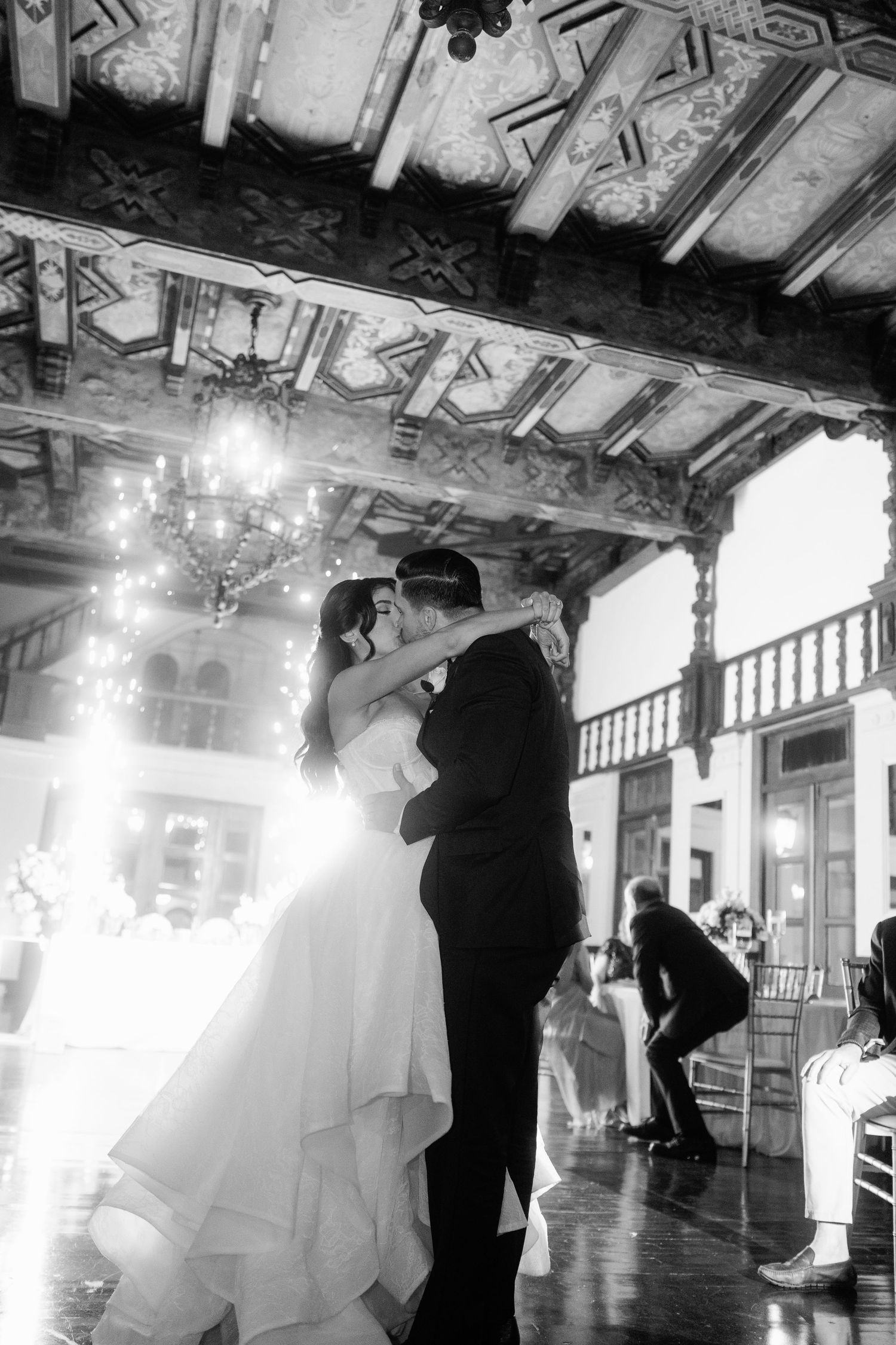 Black and white photo of bride and groom sharing first dance in grand ballroom with chandelier.