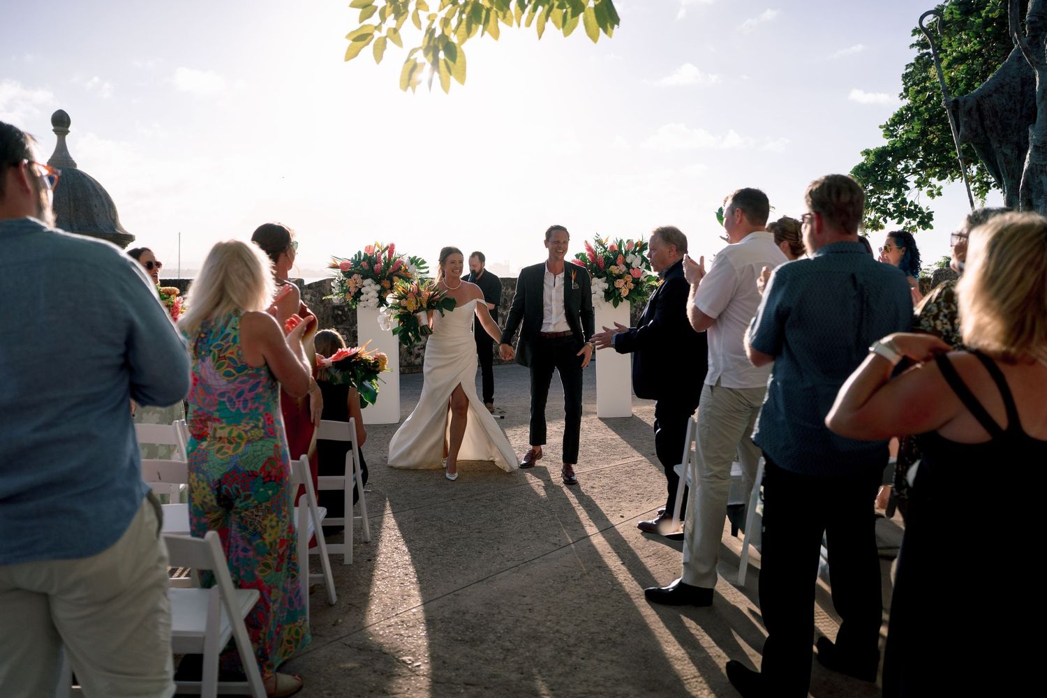 Bride and groom walk down the aisle at an outdoor wedding ceremony with guests standing on both sides.