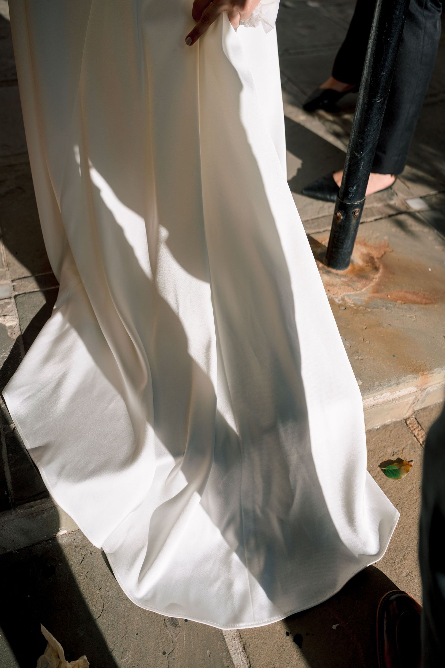 Bride holding flowing white wedding dress train in sunlight on outdoor stone steps.