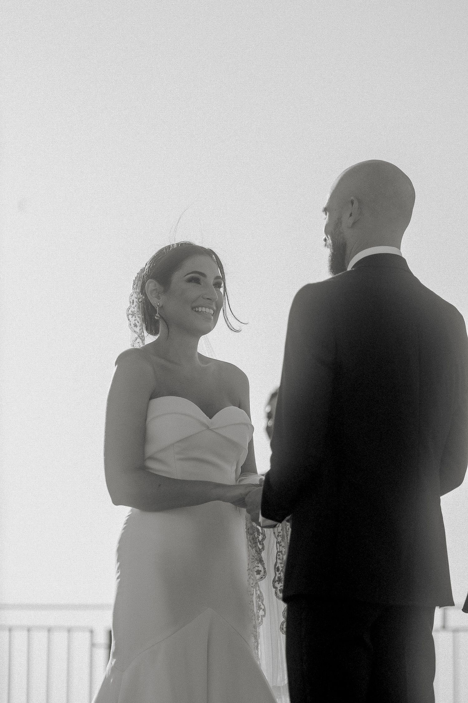Black and white photo of a smiling bride in a strapless gown holding hands with a groom in a suit at their wedding ceremony.