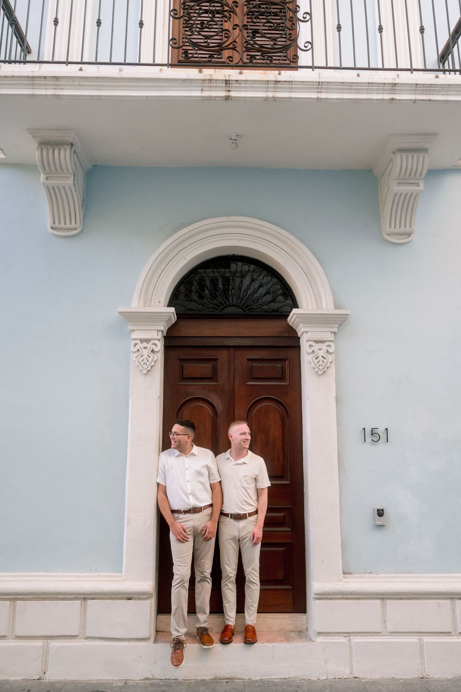Two men in matching beige outfits stand before an elegant blue building with ornate wooden door numbered 151.