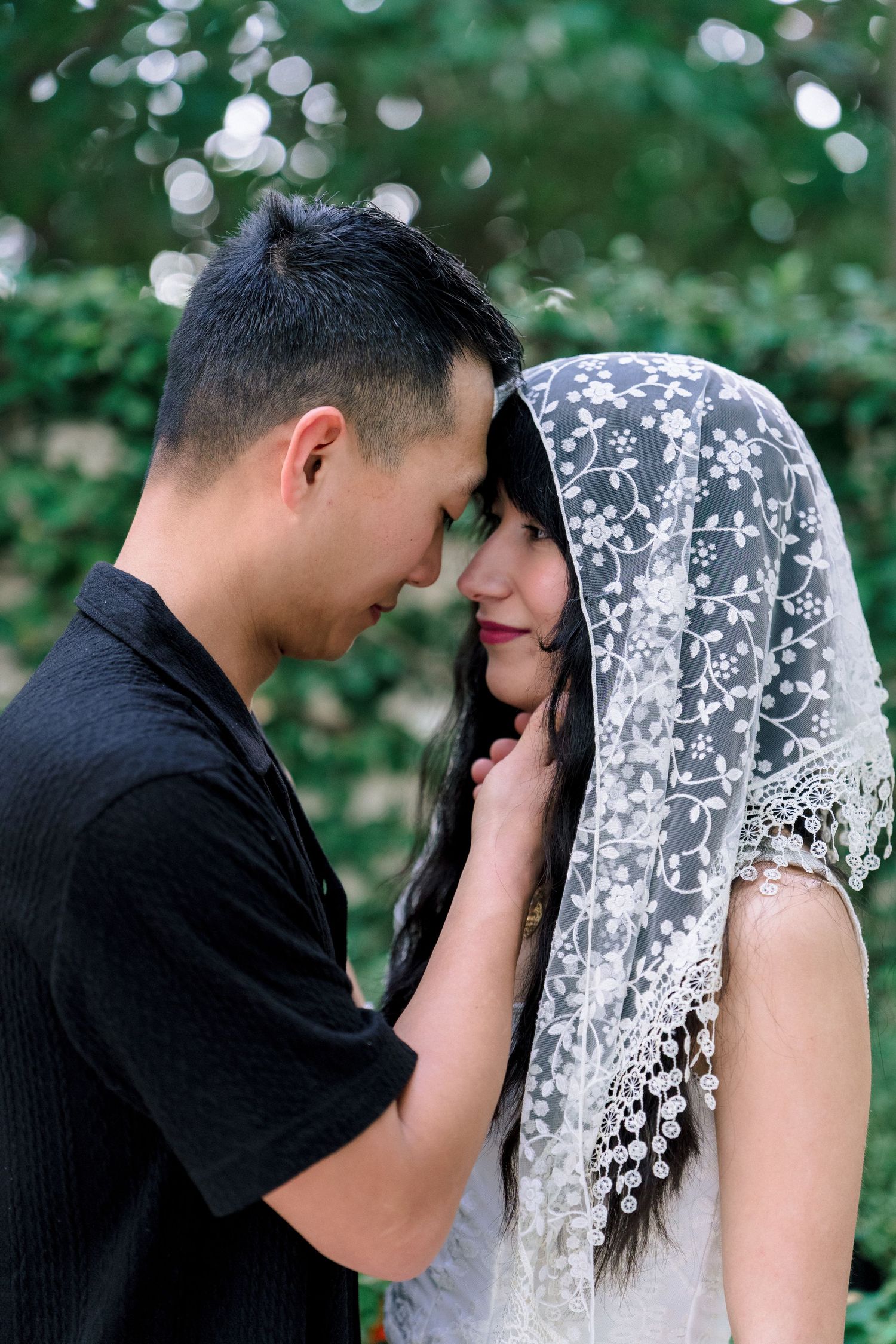 Romantic Asian couple in wedding attire share an intimate moment outdoors, bride wearing elegant lace veil.