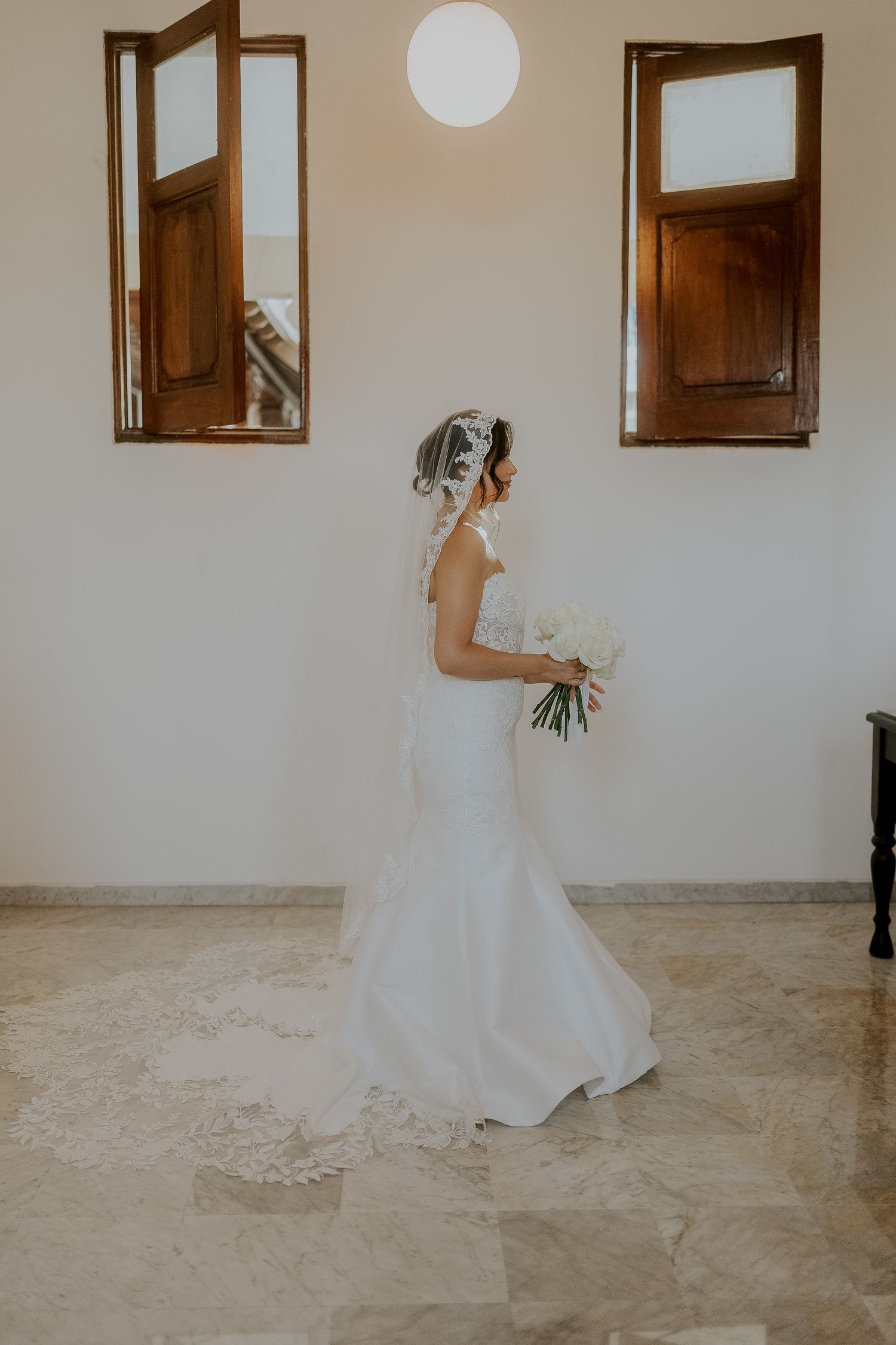 Elegant bride in fitted white gown with lace veil holding white bouquet inside a bright venue.