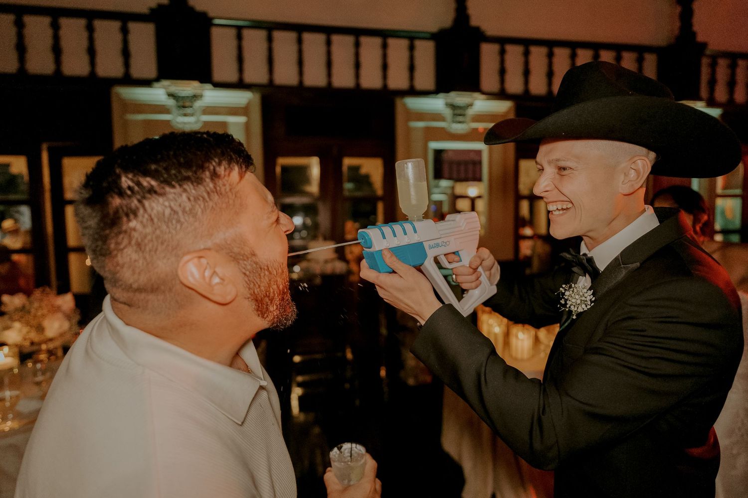 Groom in cowboy hat playfully squirts drink into guest's mouth at lively wedding reception.