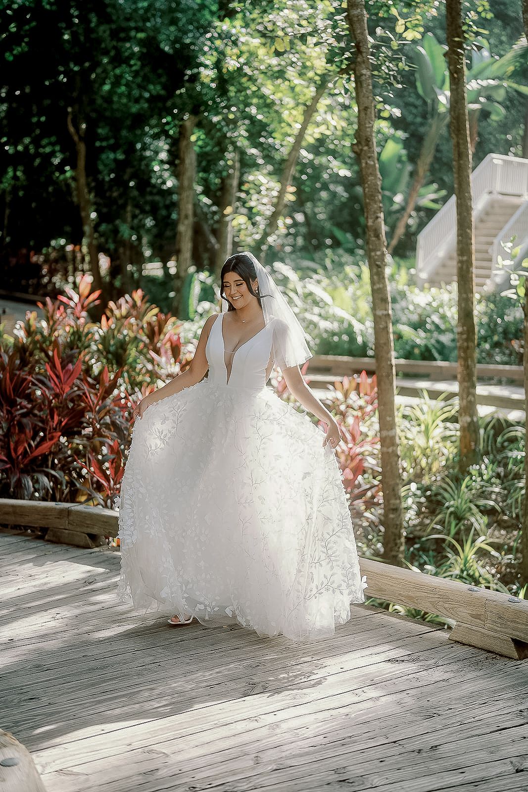 Bride in a white ballgown wedding dress with veil stands on a wooden deck surrounded by lush tropical greenery.