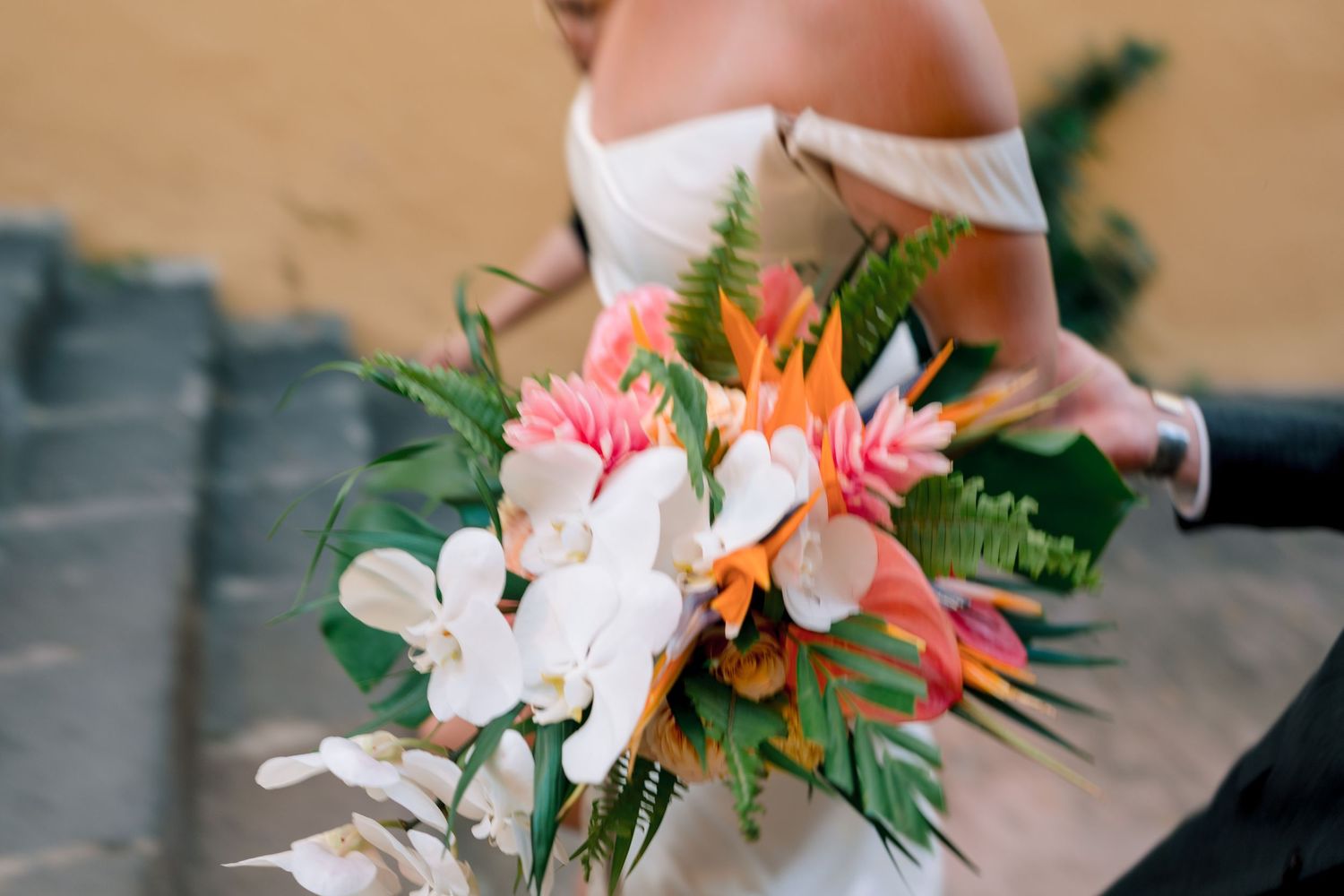 Bride holds a vibrant tropical wedding bouquet with orange, pink, and white flowers and lush green foliage.