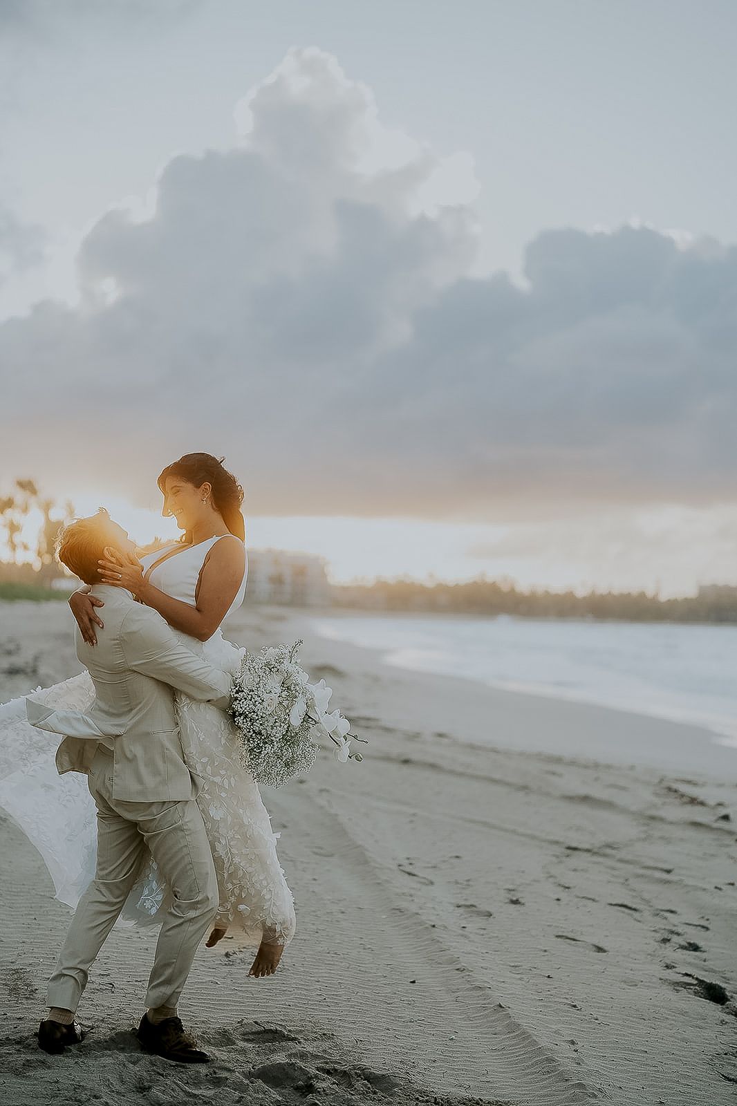 Groom lifting bride on beach at golden sunset with bouquet and veil.
