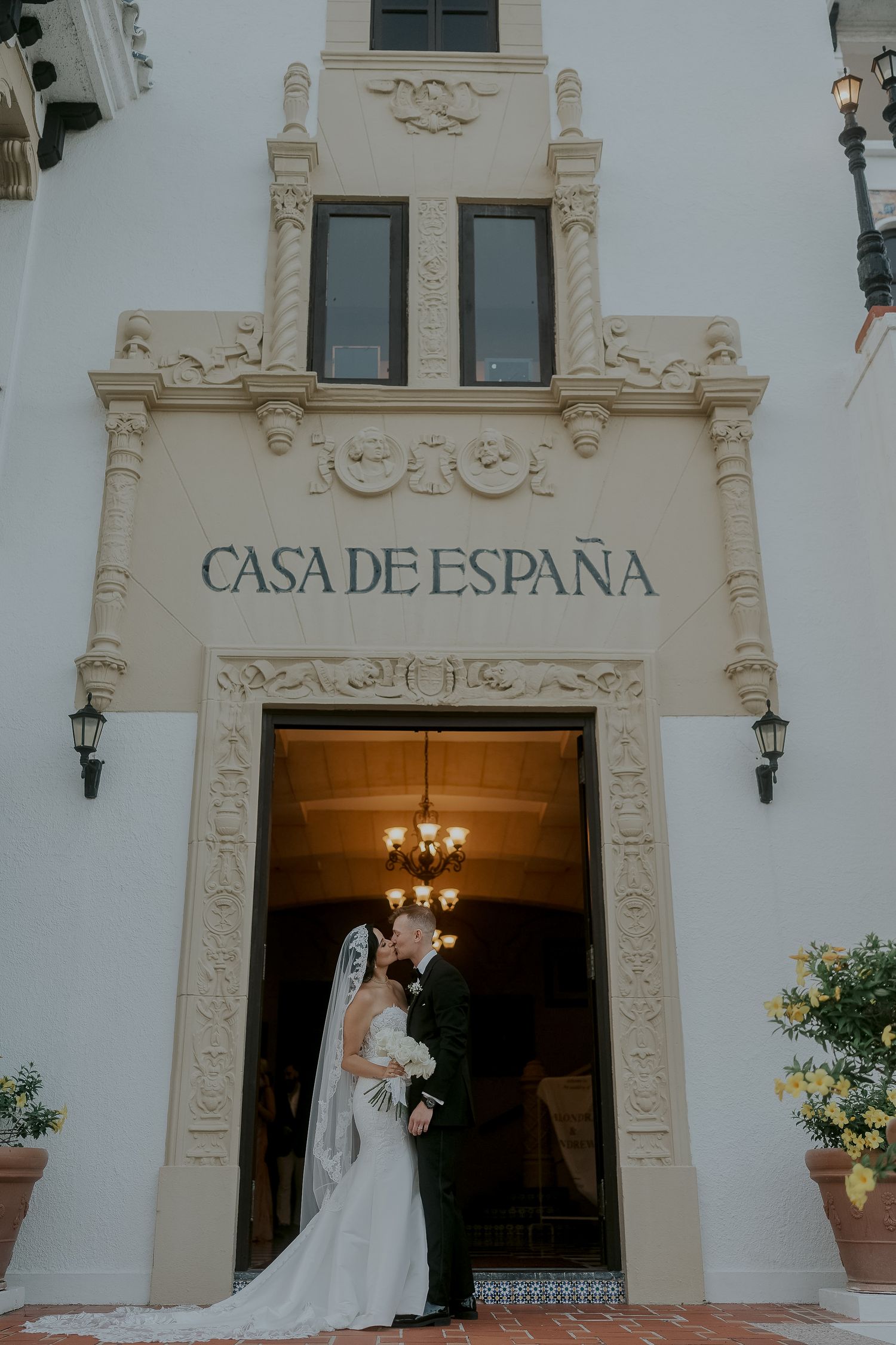 Wedding couple kissing under ornate Casa de Espana building entrance with chandelier and decorative facade.