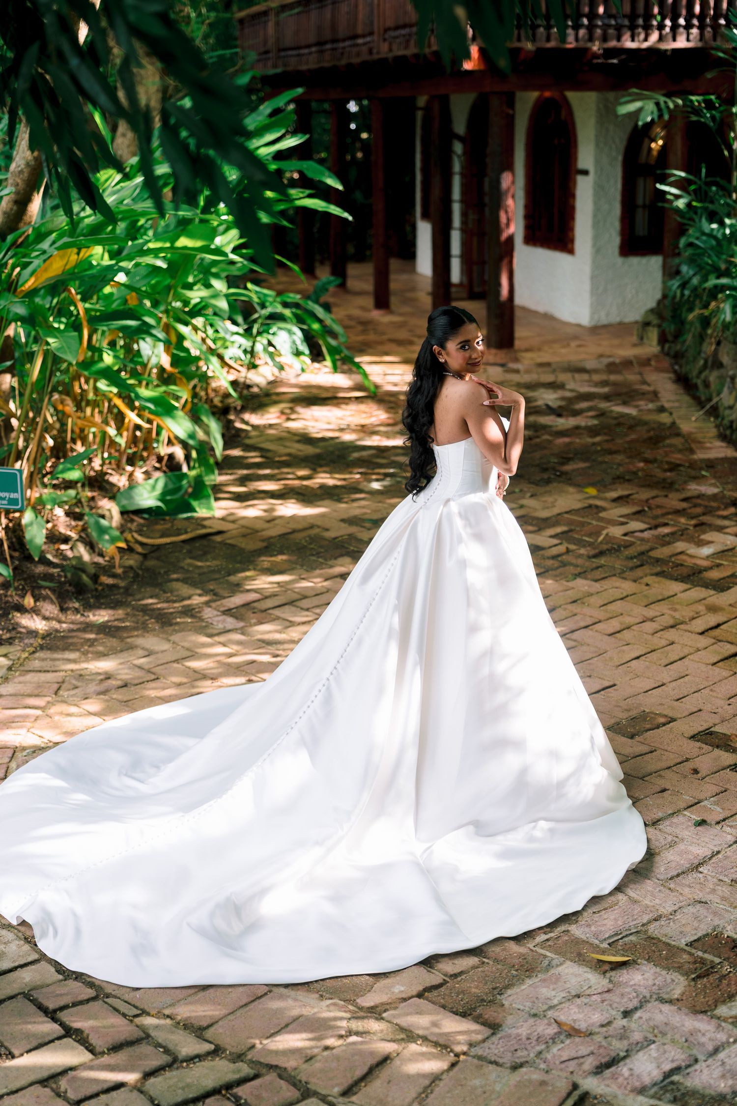 Bride in strapless white ball gown with long train stands on brick path surrounded by tropical greenery.