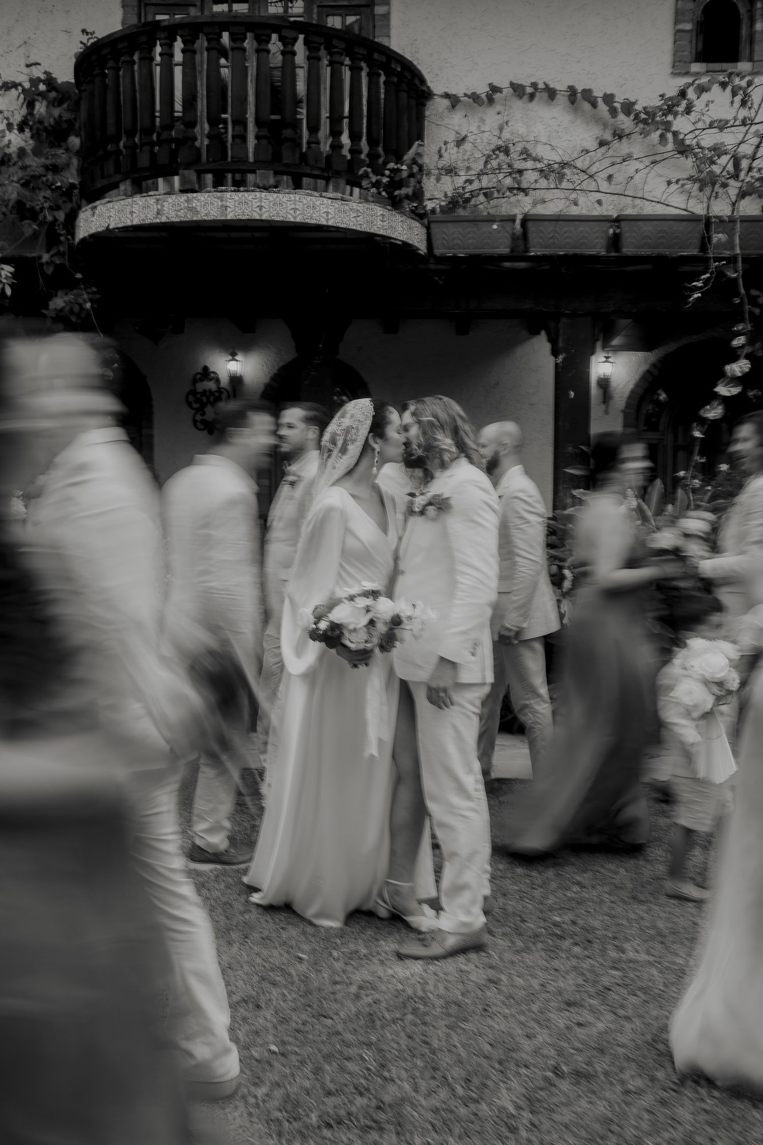 Black and white wedding photo of couple kissing amid blurred guests outside a rustic venue.