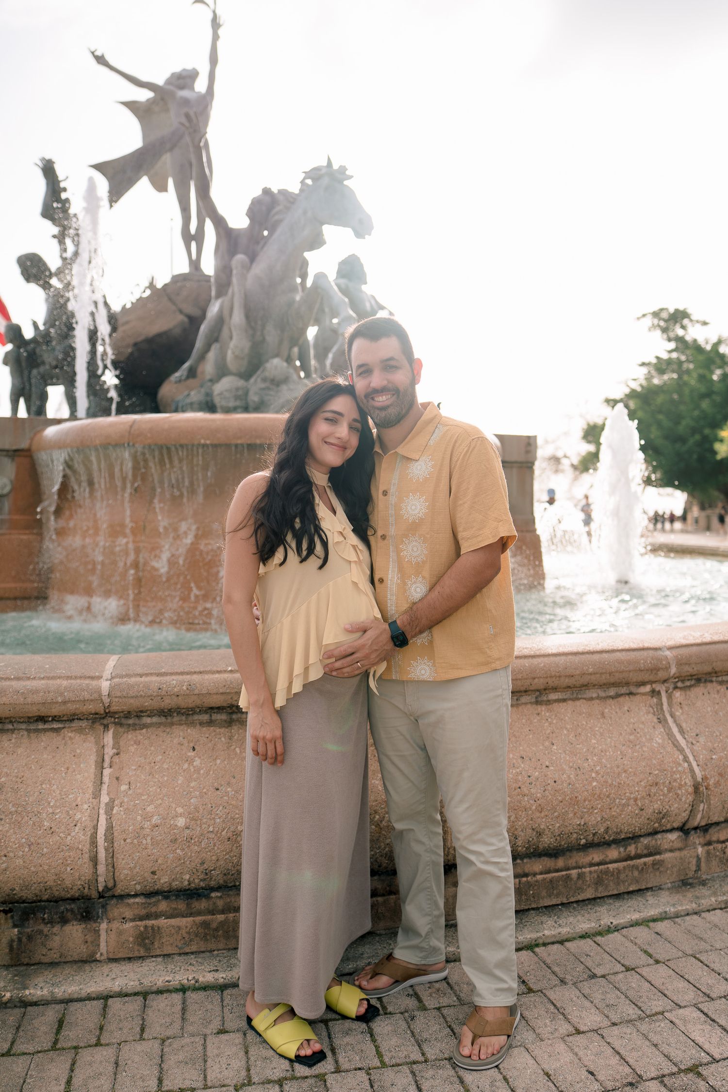 Pregnant couple posing together in front of an ornate fountain sculpture, woman in maxi dress, man in yellow shirt.