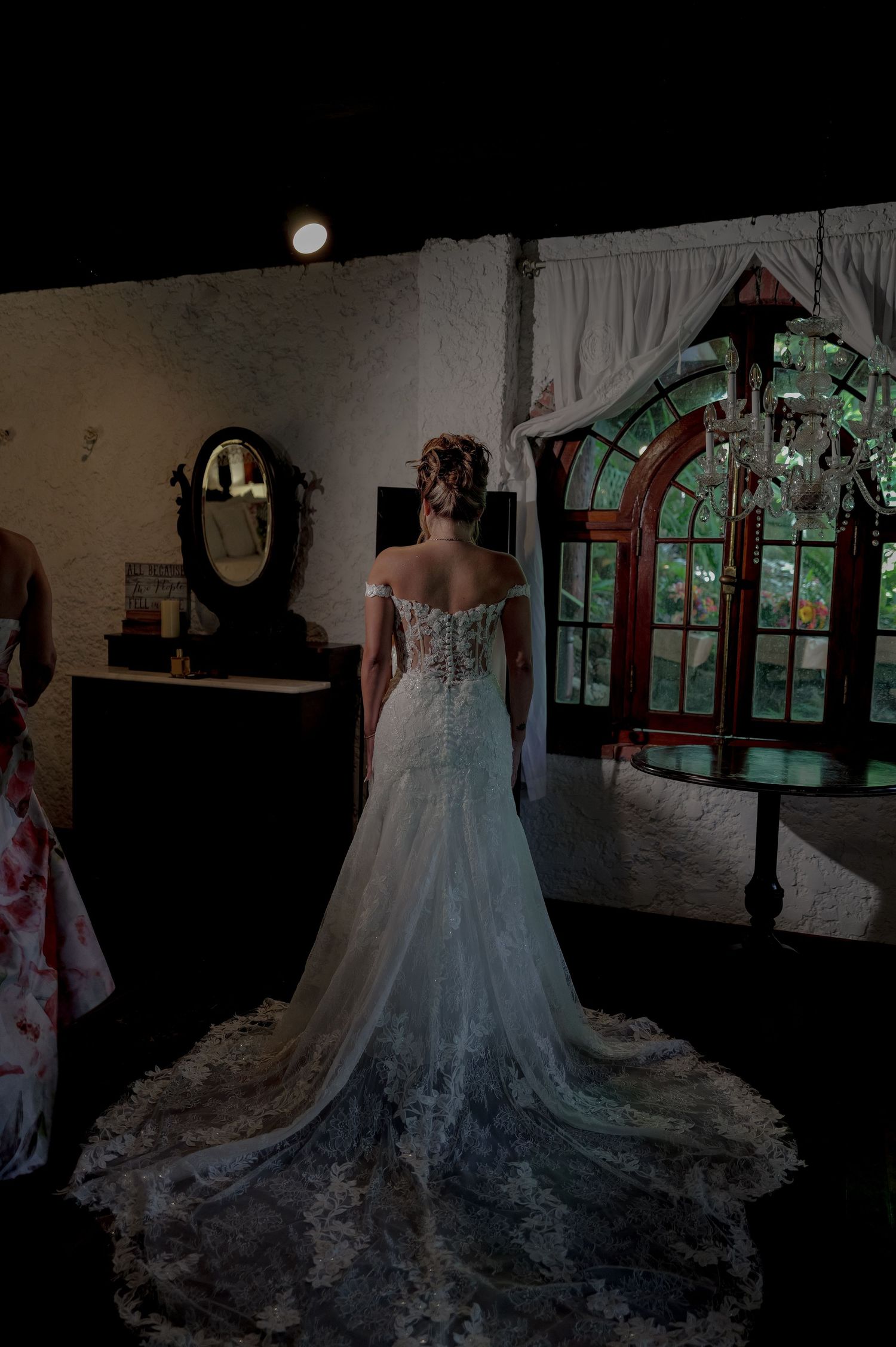Bride in elegant lace off-shoulder wedding gown with long train standing before ornate stained glass window.