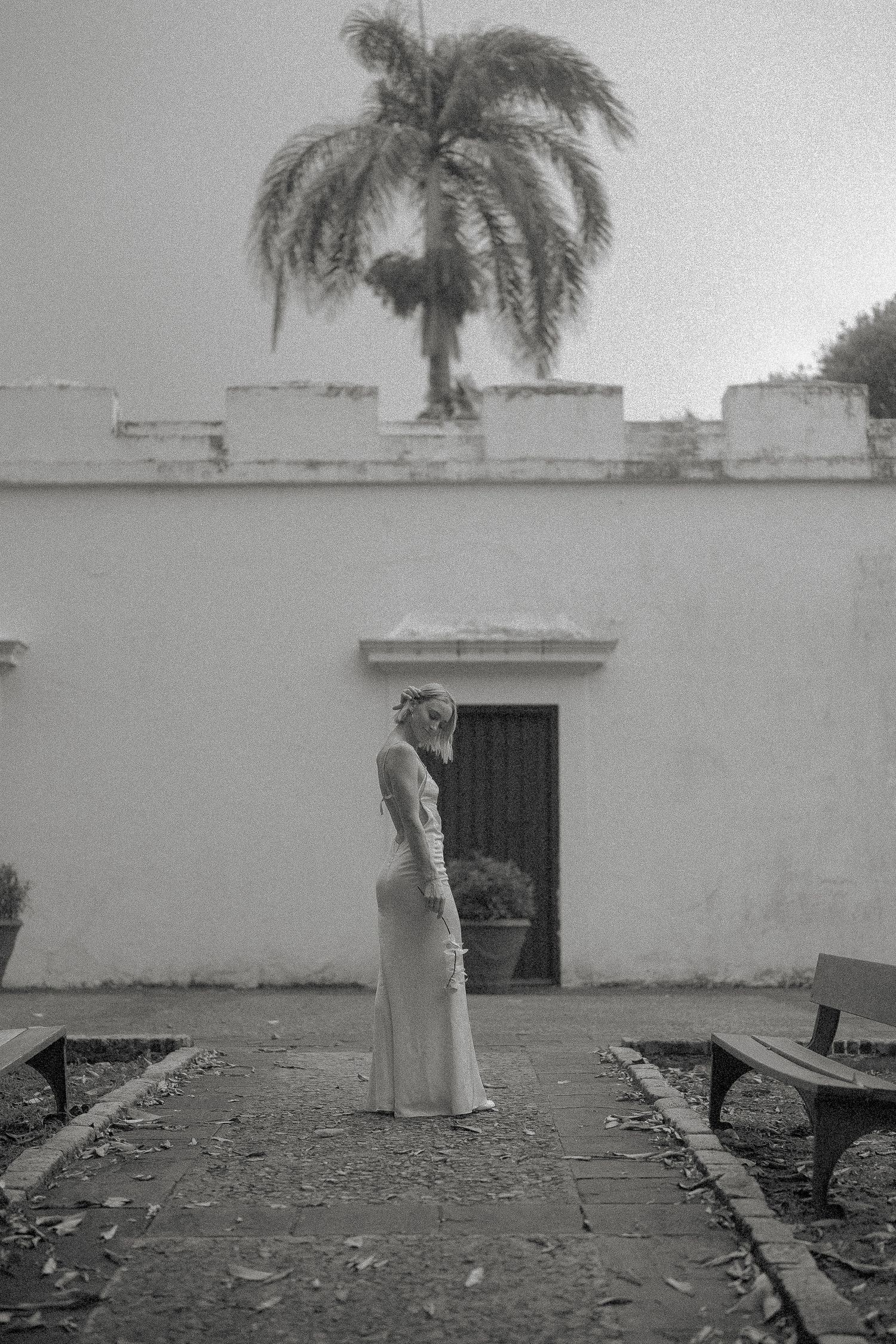 Bride in white gown standing before historic white building with palm tree in black and white photo.
