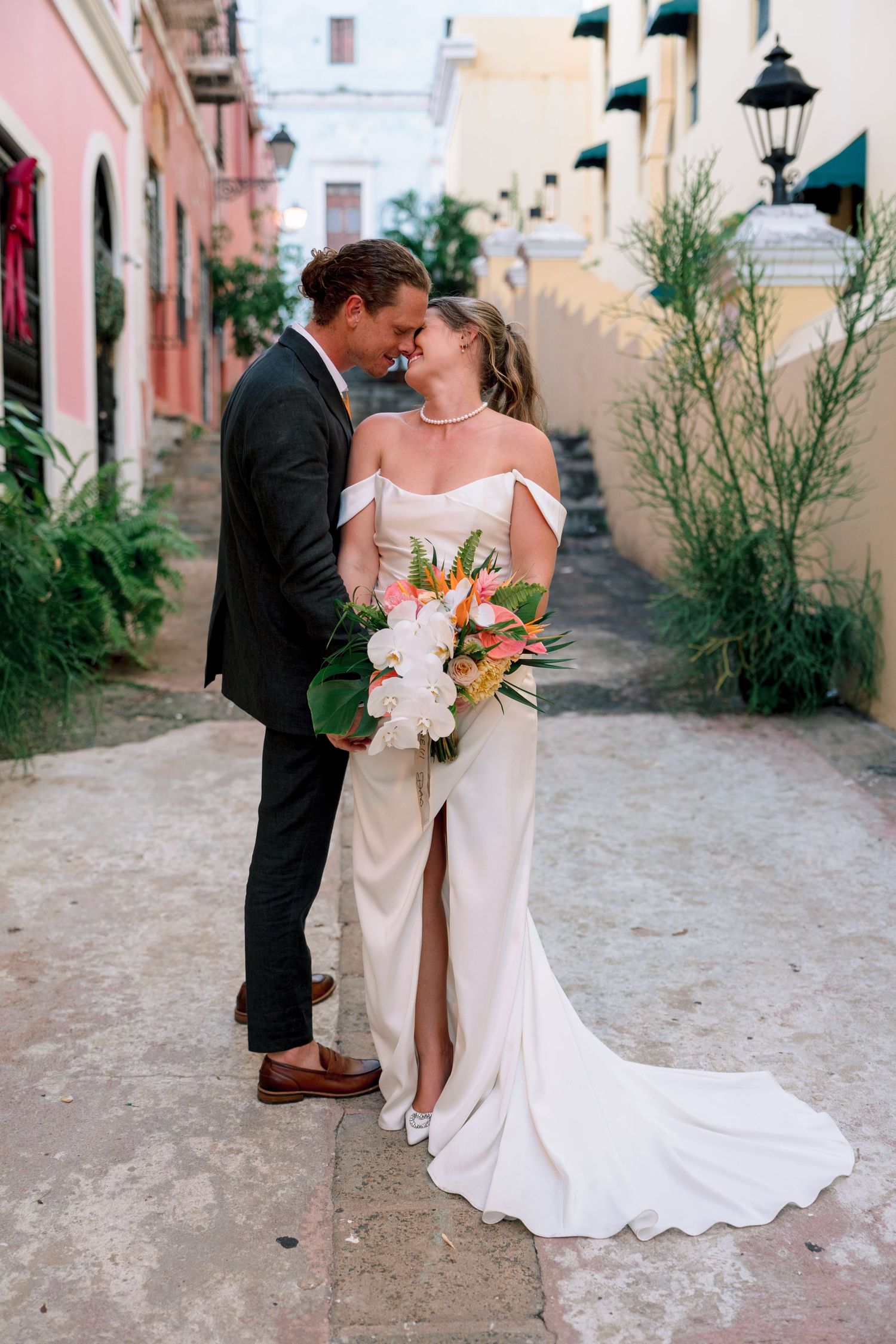Bride and groom share a romantic moment in a colorful cobblestone alley, bride holding tropical bouquet.