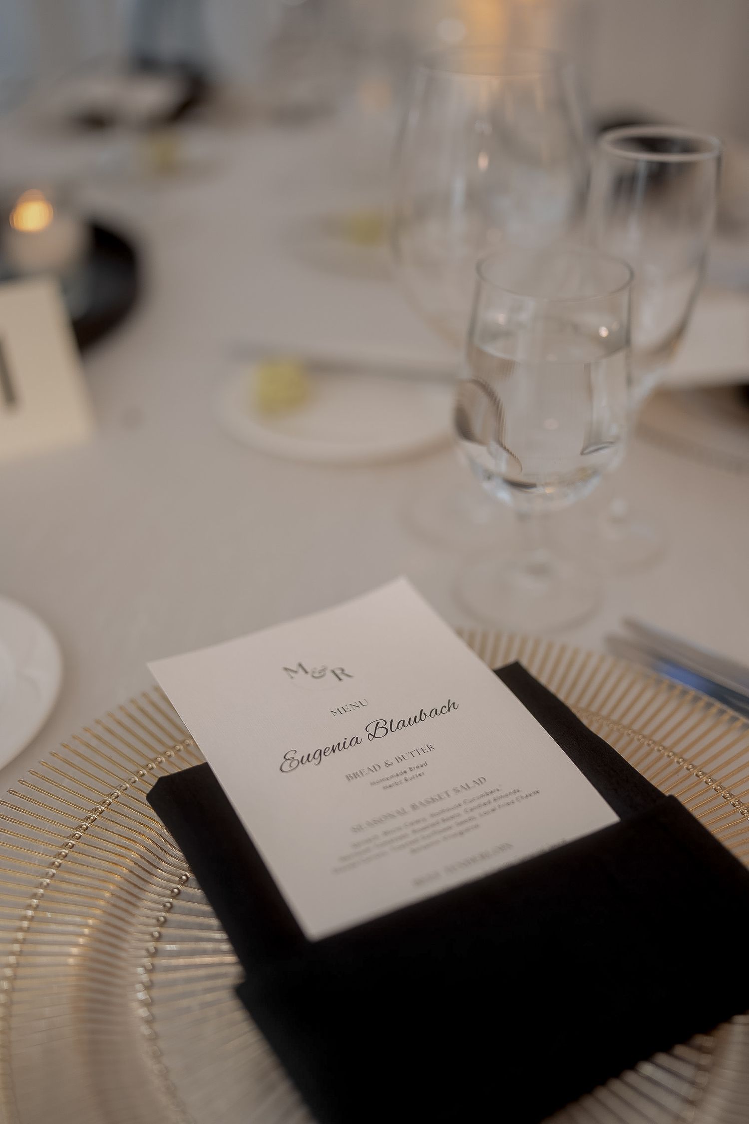 Elegant wedding place setting with personalized menu card on black napkin, gold charger plate, and crystal glassware.