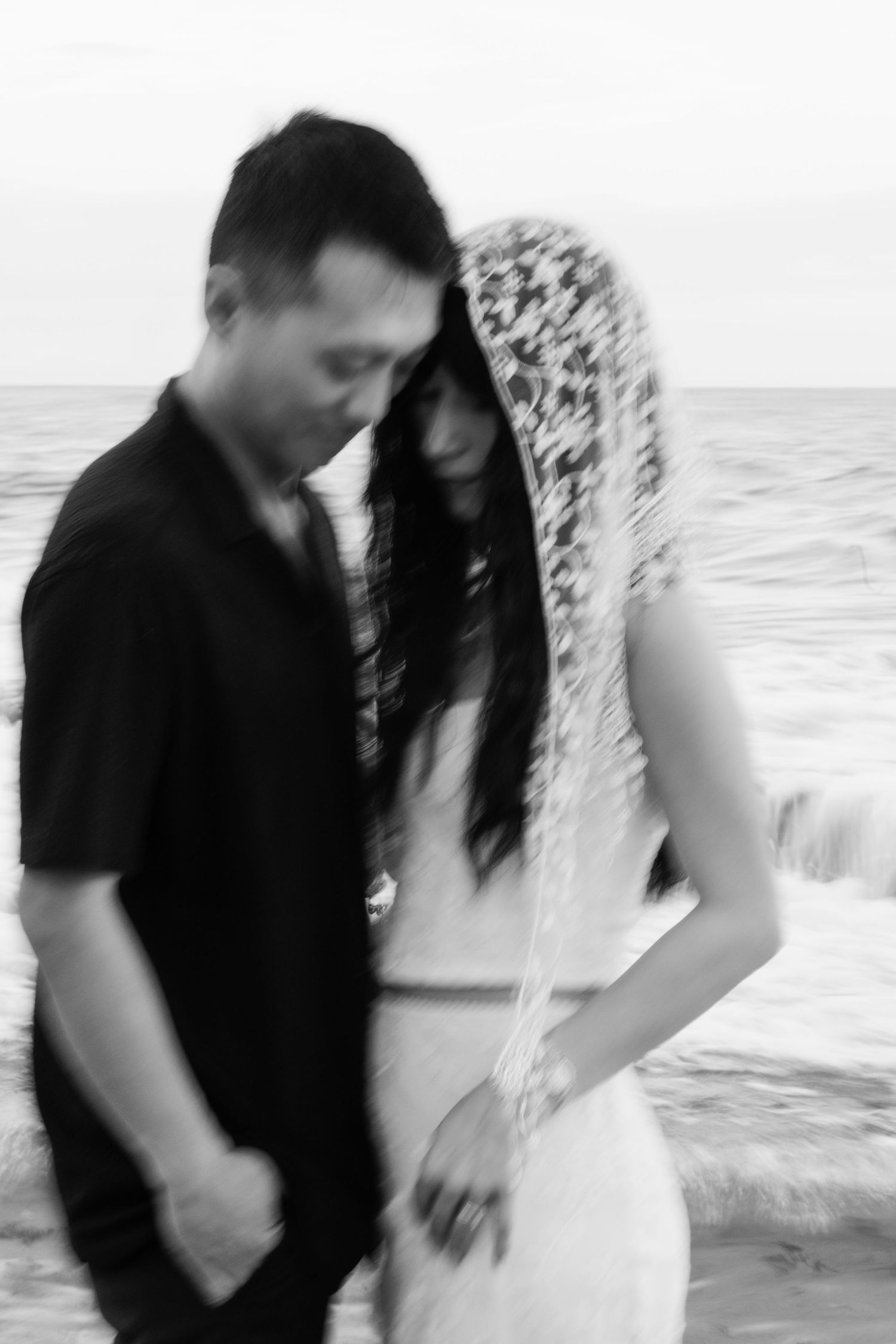 Black and white photo of a couple embracing near ocean waves, bride wearing a lace veil in a romantic blur effect.