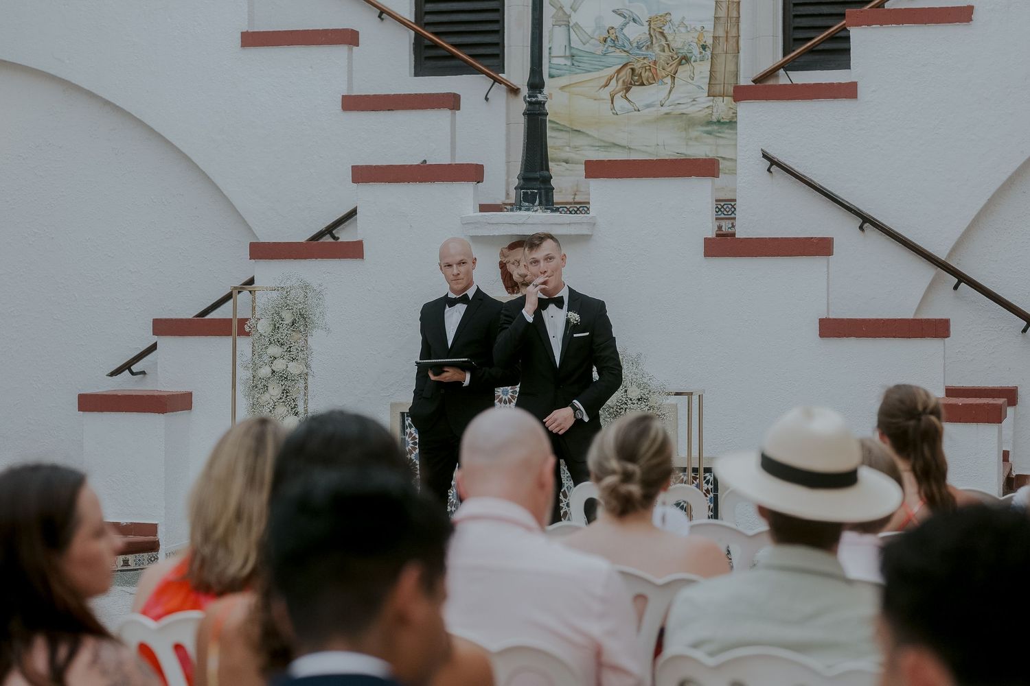 Two grooms in tuxedos stand at outdoor wedding ceremony altar before seated guests at elegant white venue.