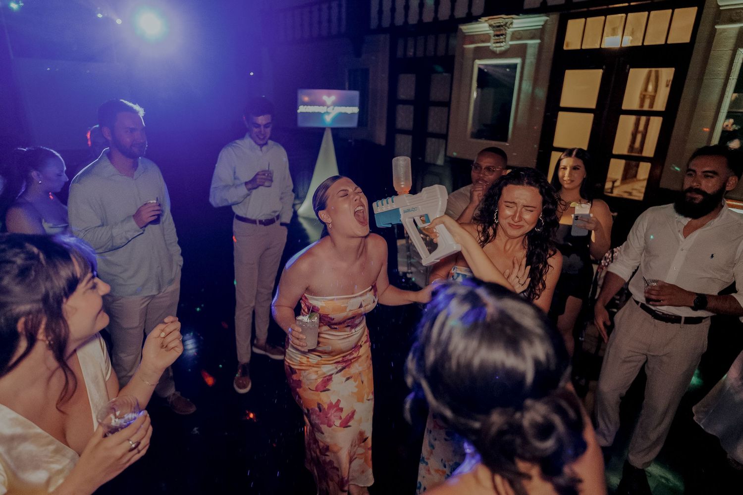 Wedding guests dancing and celebrating under colorful lights at lively indoor reception party.