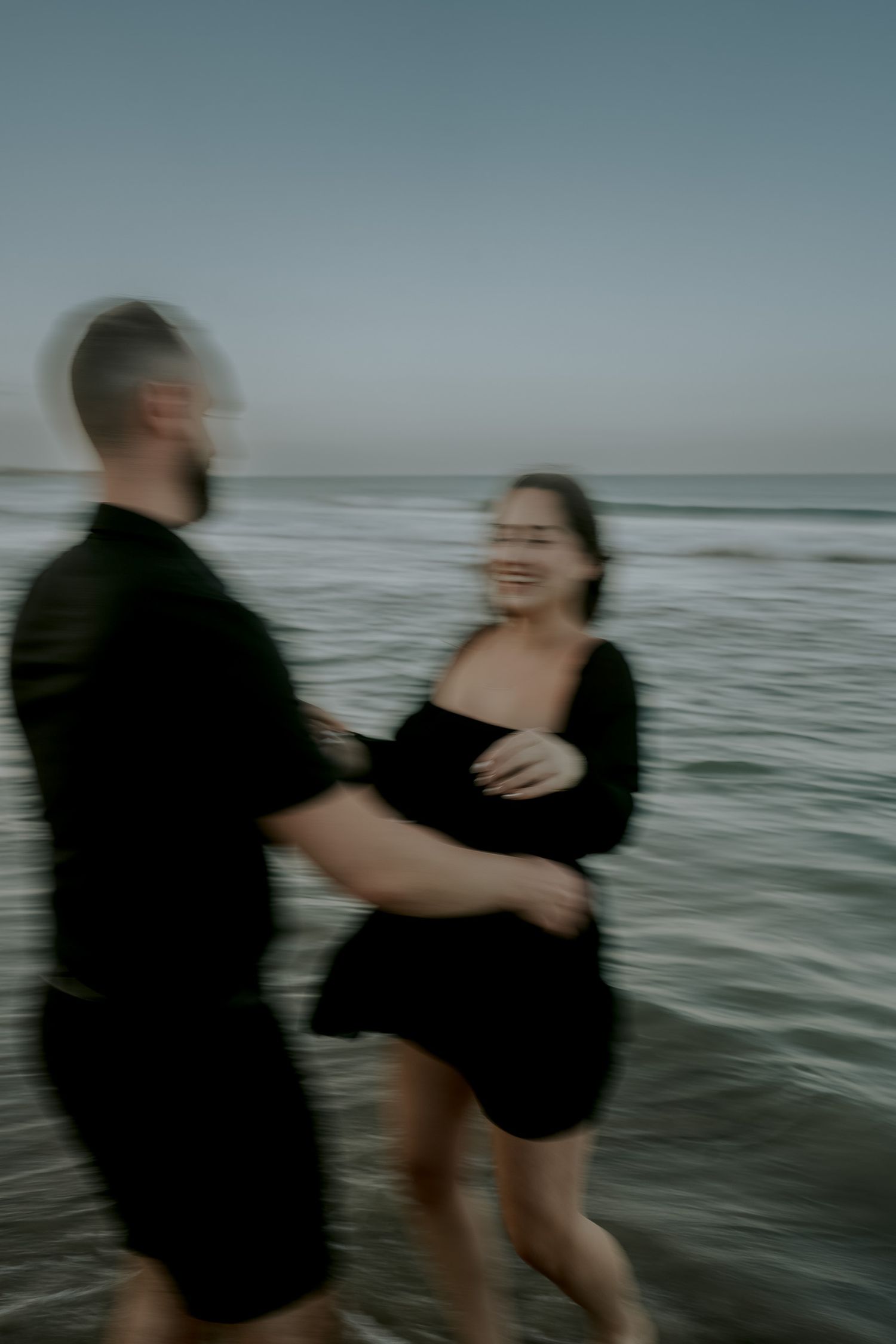 Blurry motion shot of a couple in black outfits laughing and running together on a beach at dusk.