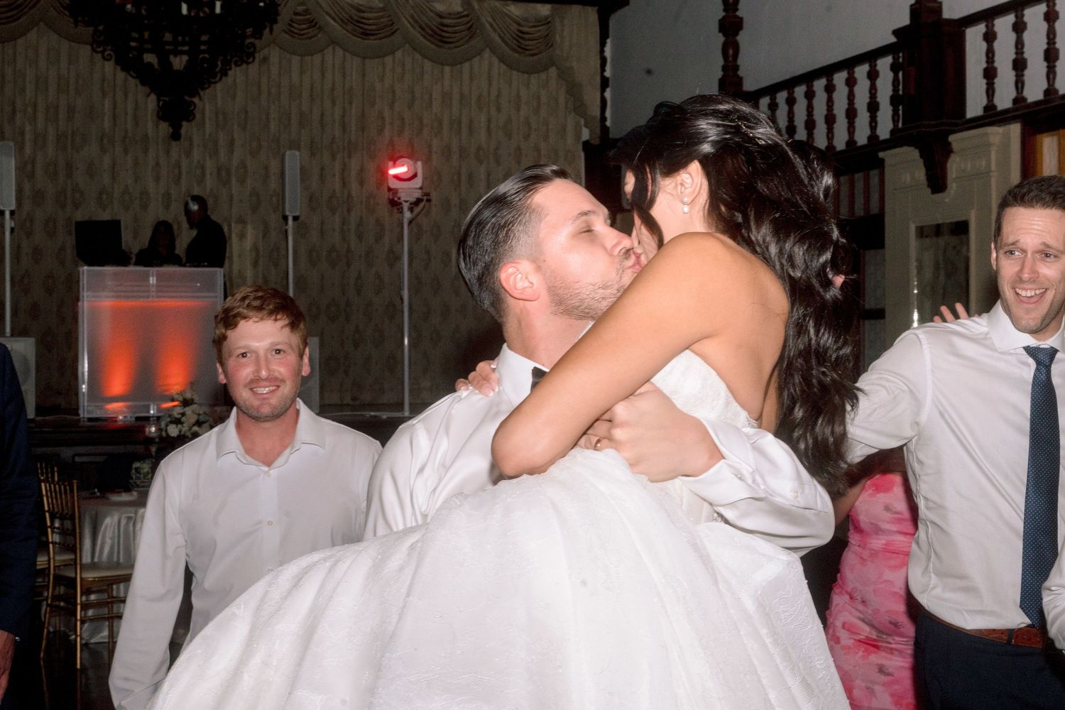 Groom lifting bride in white dress during wedding reception dance while guests smile in background.