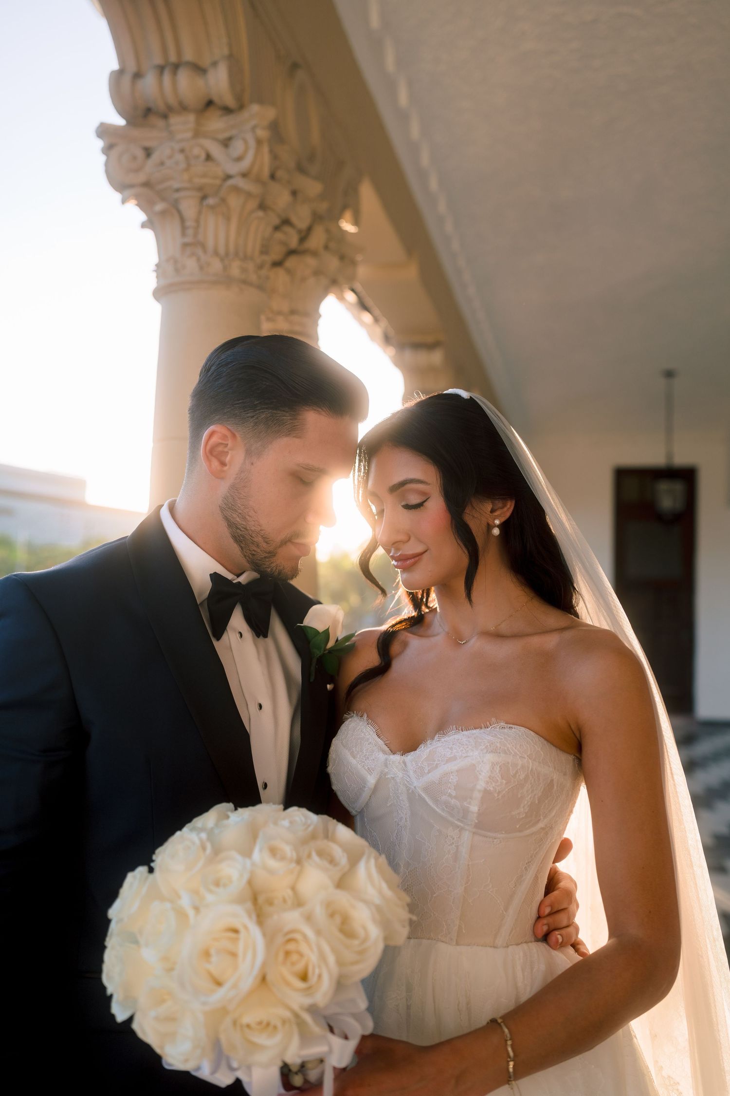 Bride and groom share intimate moment on sunlit colonnade, she holds white rose bouquet.