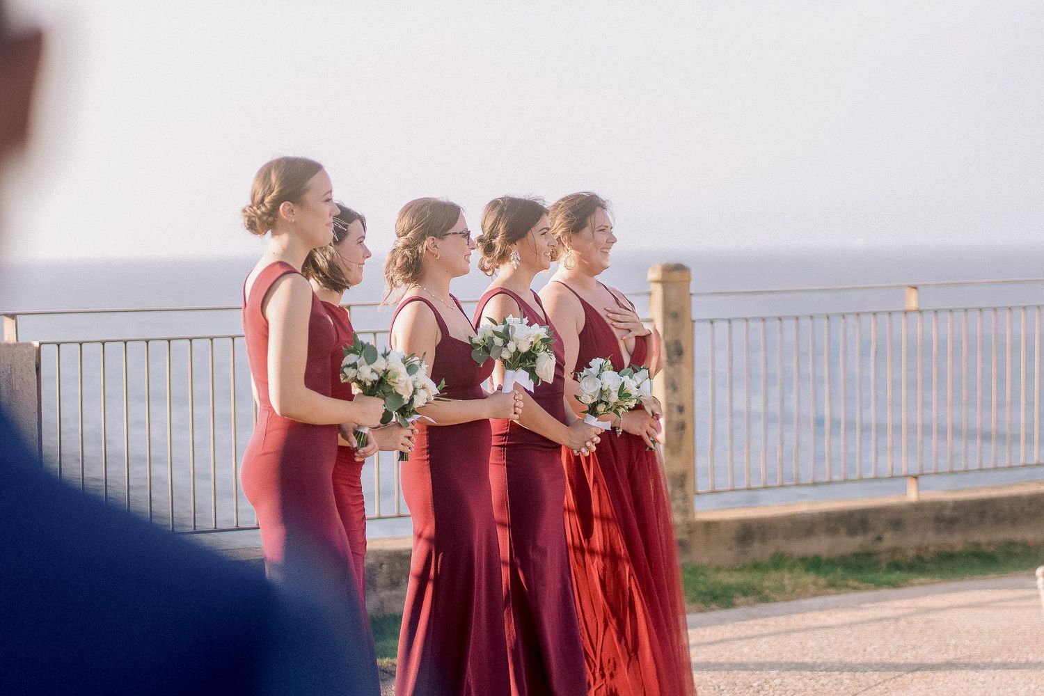 Four bridesmaids in red dresses holding white bouquets standing together at outdoor coastal wedding venue.