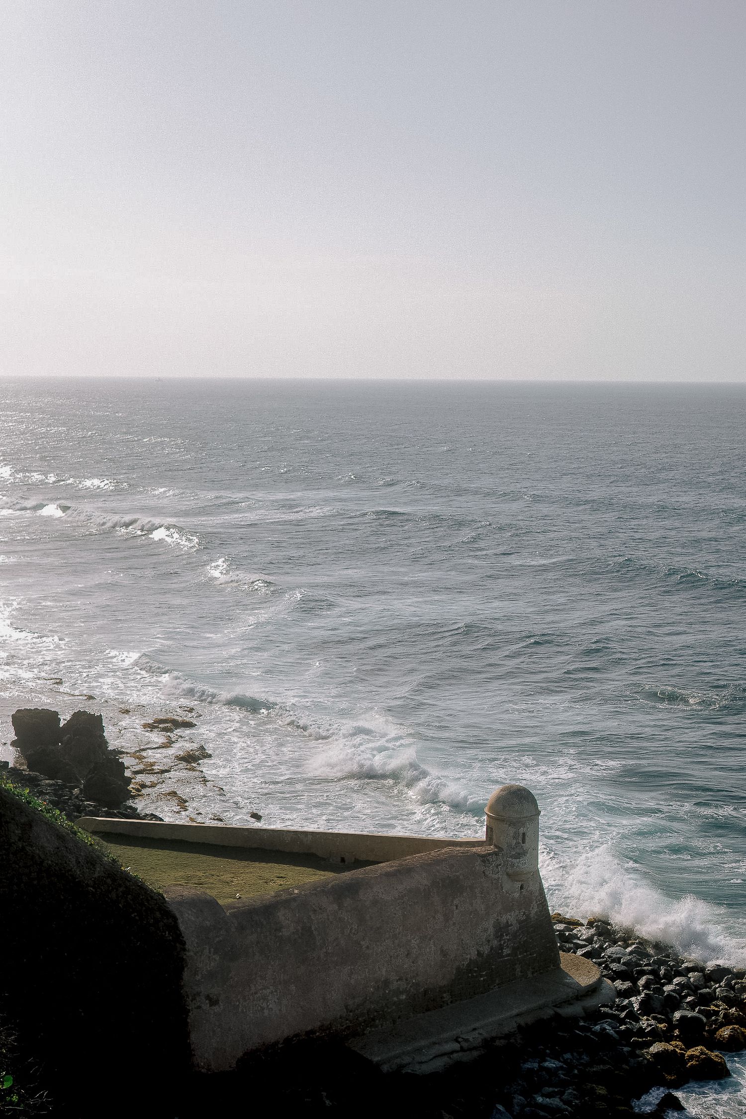 Stone fortress wall overlooks crashing ocean waves under a hazy sky at a coastal cliffside.