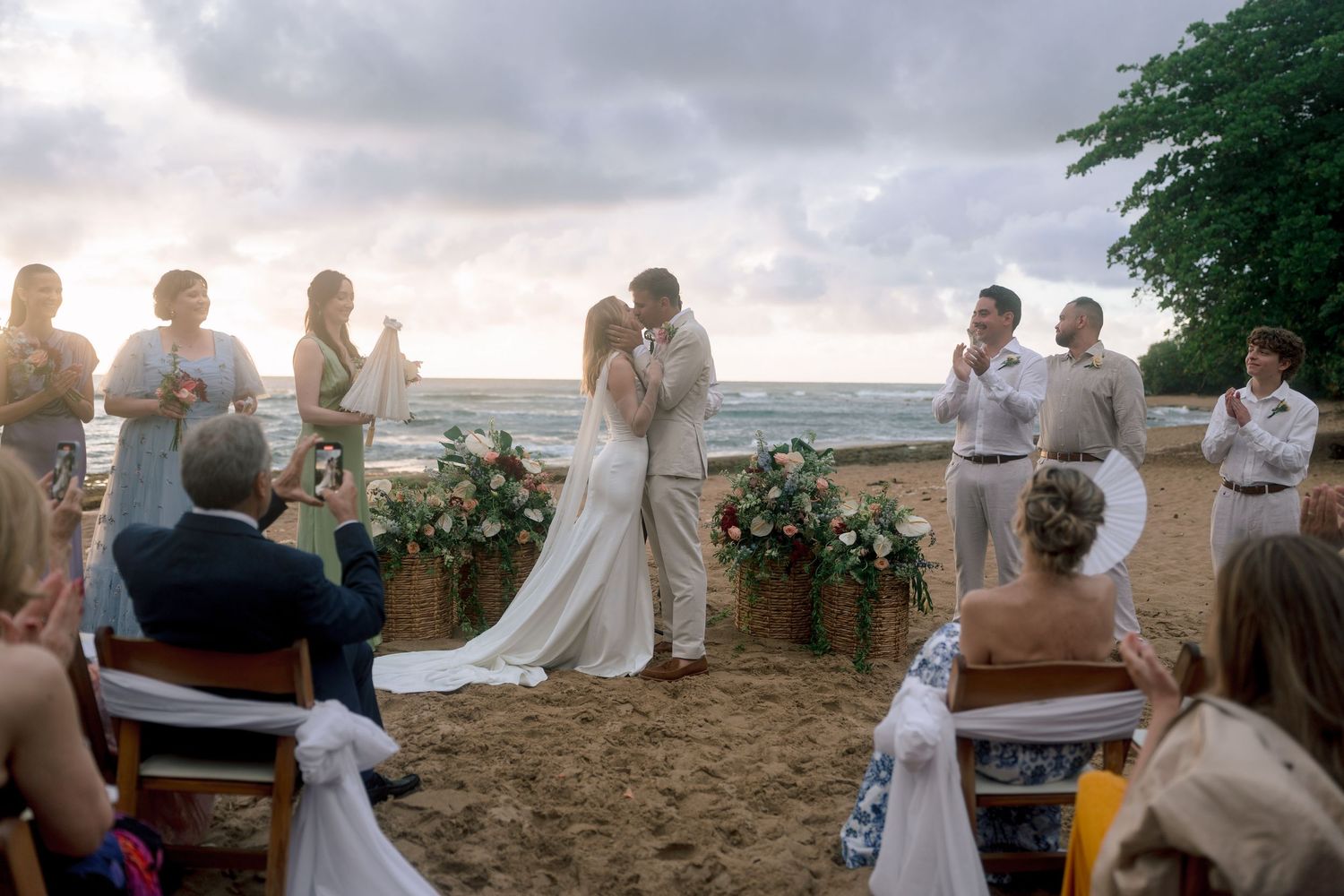 Couple shares first kiss at romantic beach wedding ceremony at sunset with guests applauding.
