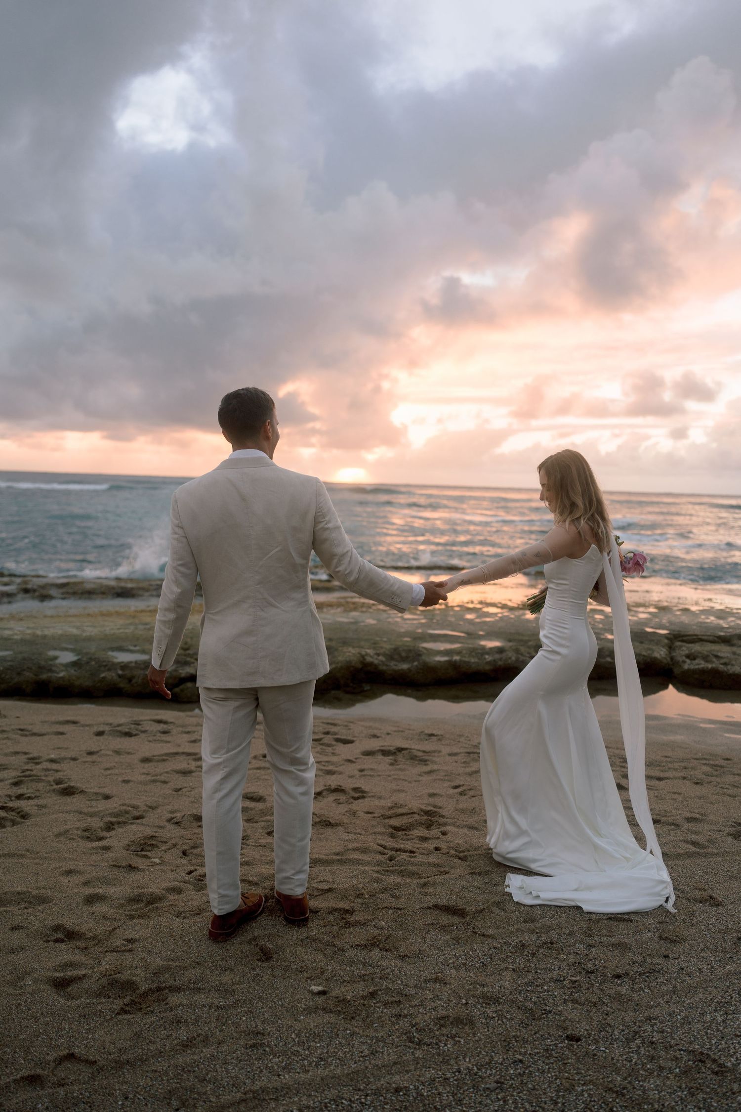 Bride and groom hold hands on a sandy beach at sunset, waves crashing behind them.