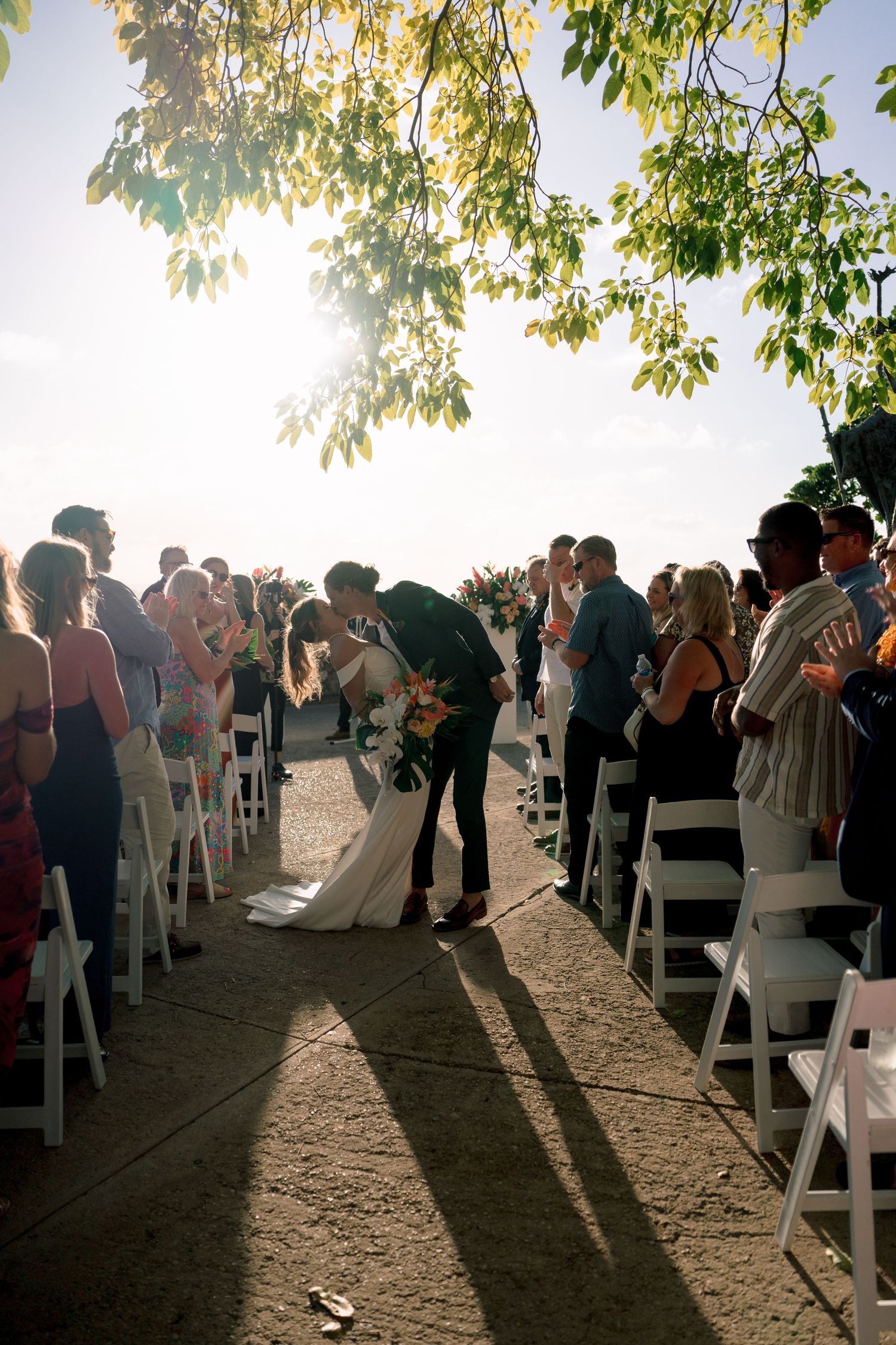 Bride and groom sharing a kiss at sunset during outdoor wedding ceremony with seated guests.