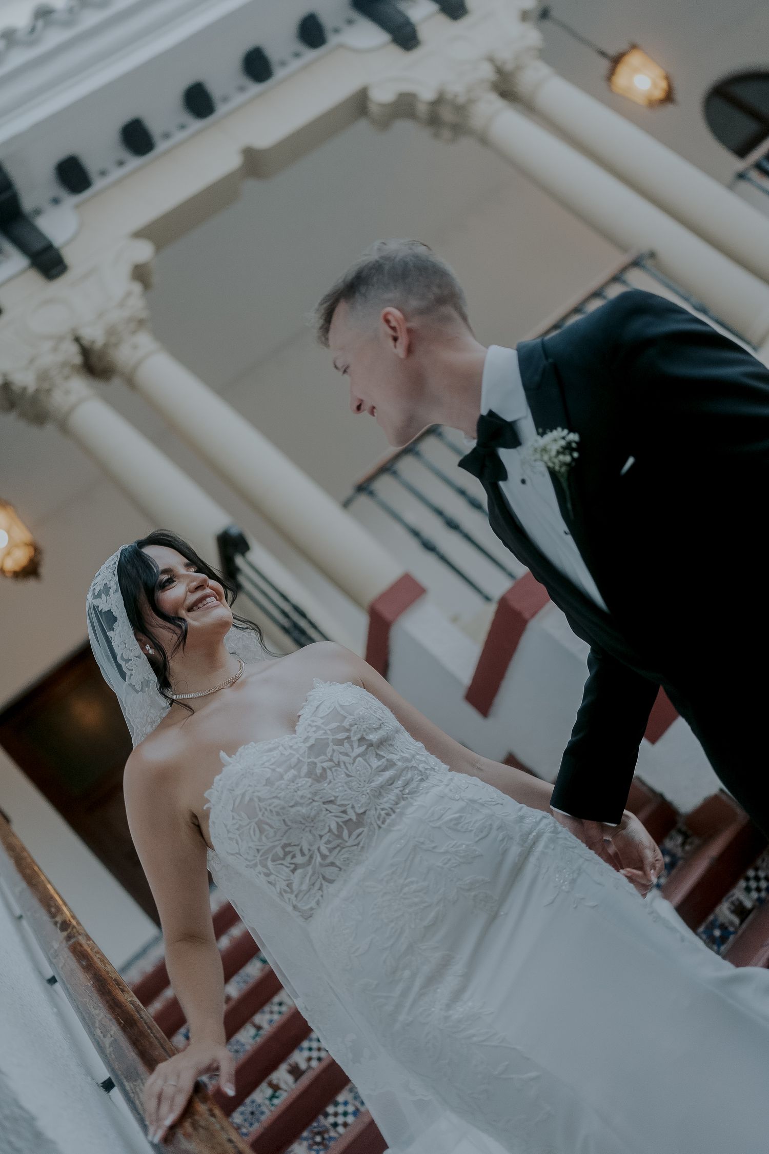Bride in lace gown smiles at groom in tuxedo on elegant staircase.