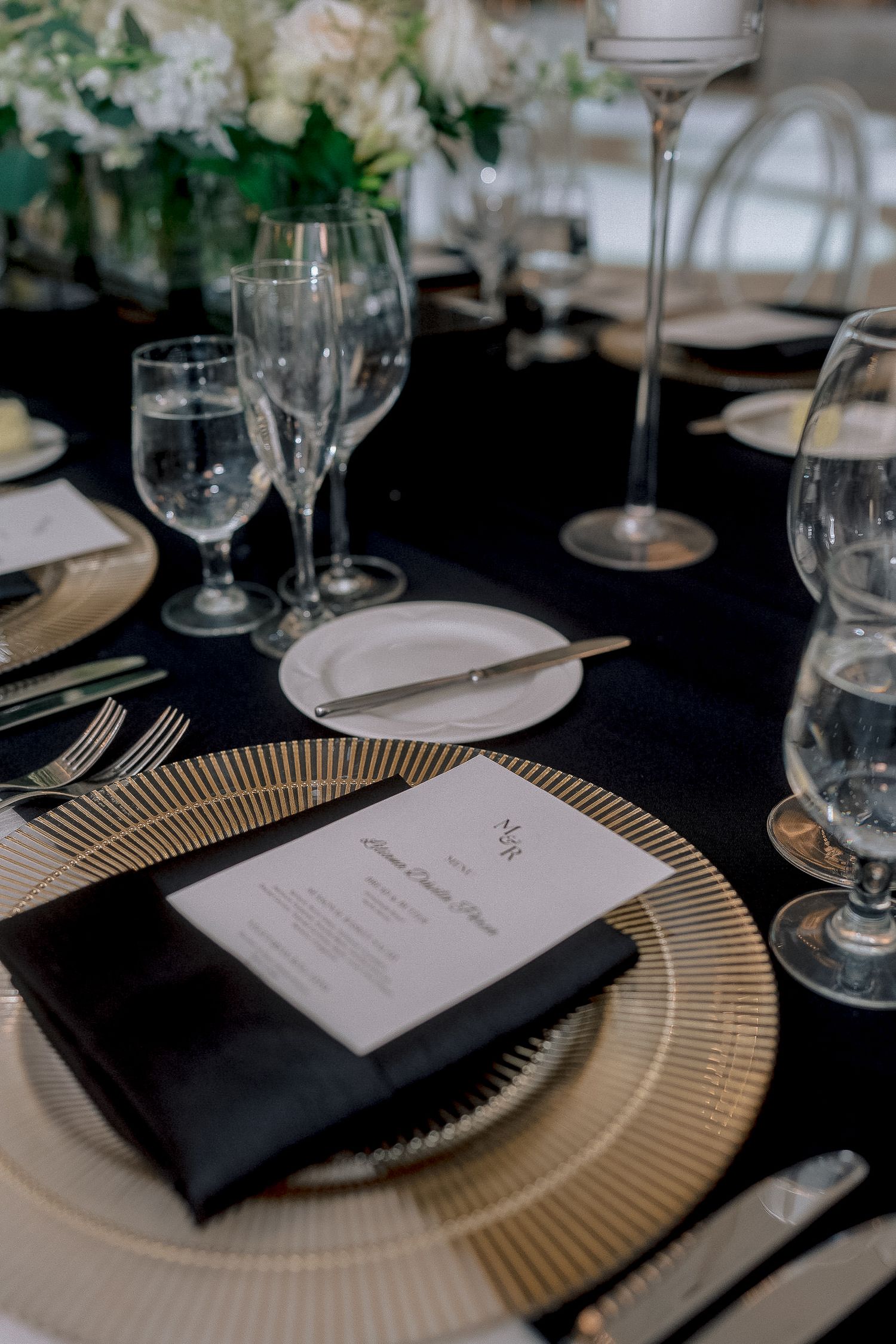 Formal wedding table setting featuring black napkin, personalized menu card, gold charger plate, and multiple wine glasses.
