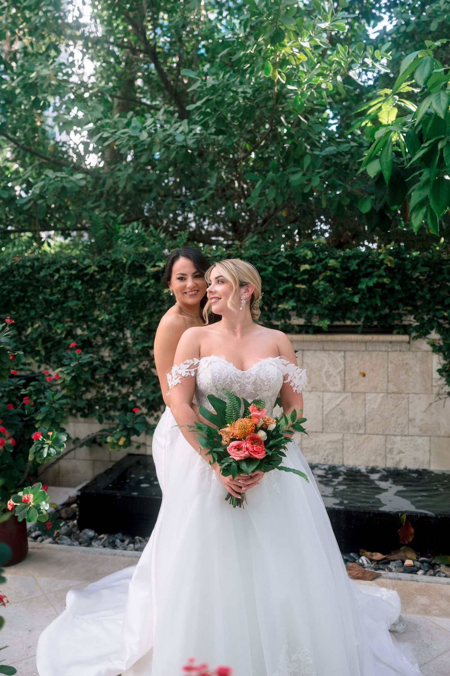 Two brides in white wedding gowns pose together in a lush garden, one holding a vibrant tropical bouquet.