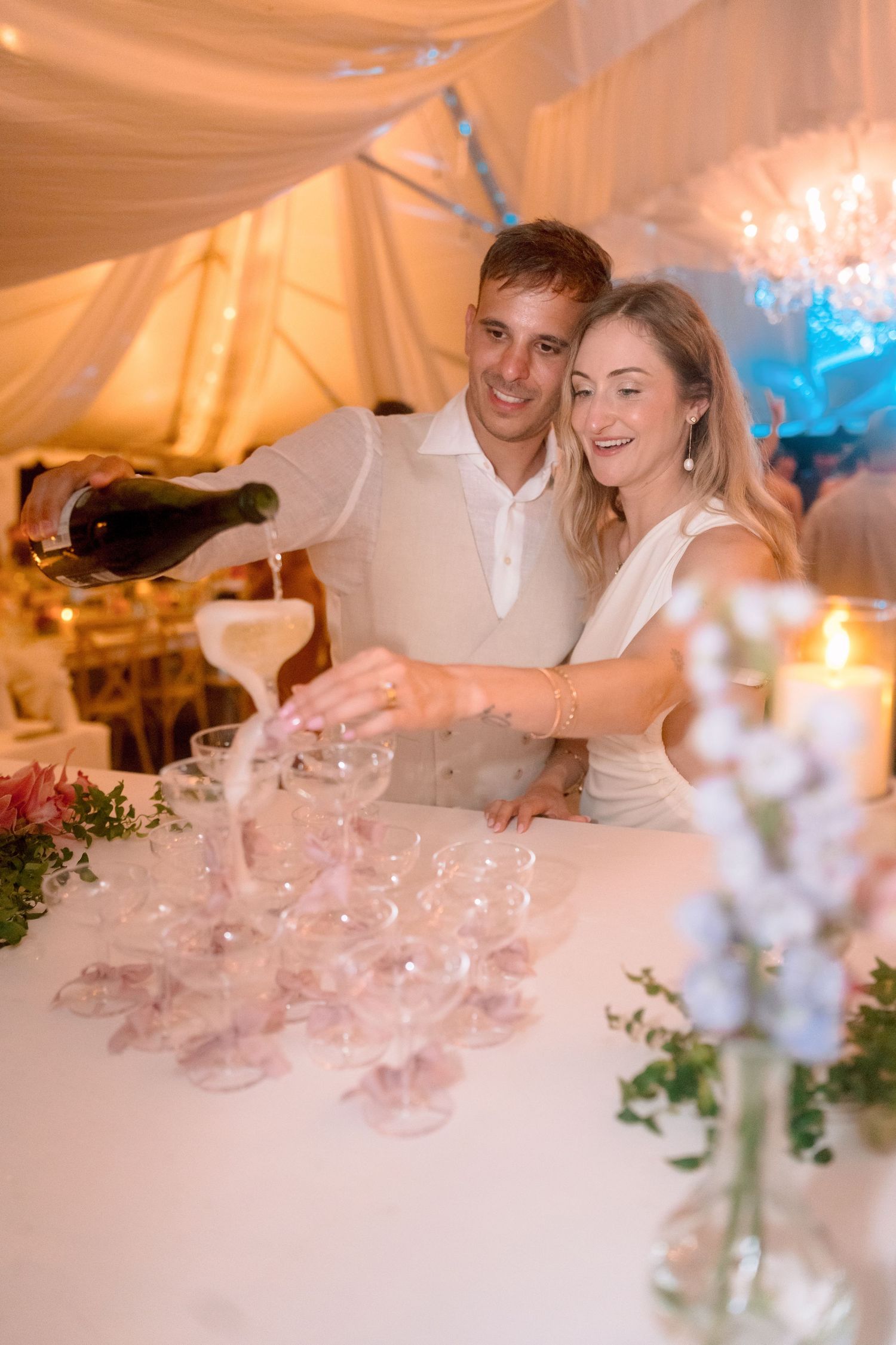 Happy couple pours champagne tower together at elegant candlelit wedding reception under white draping.