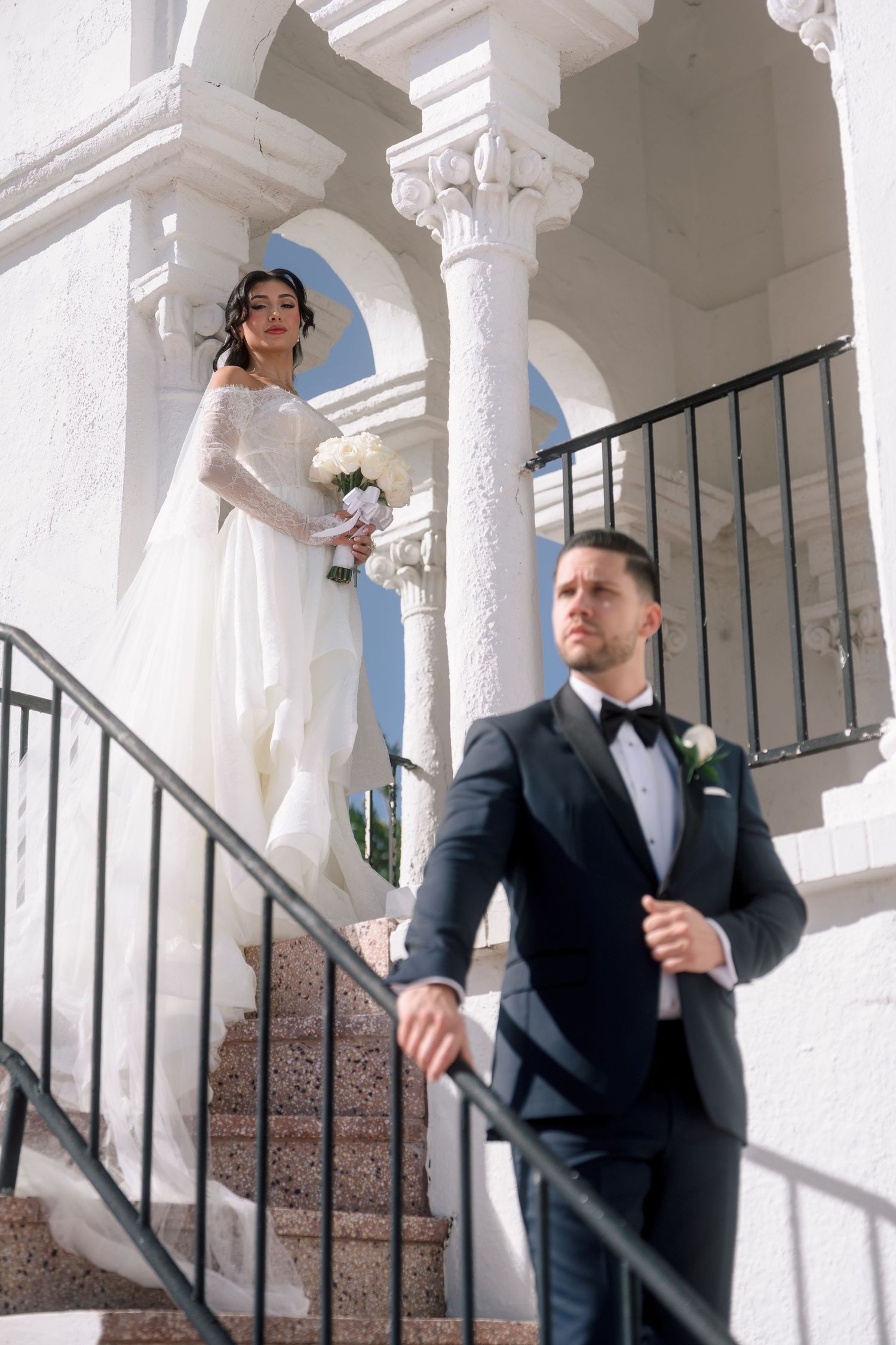 Bride in white lace gown holding bouquet stands on elegant white staircase as groom in black tuxedo descends.