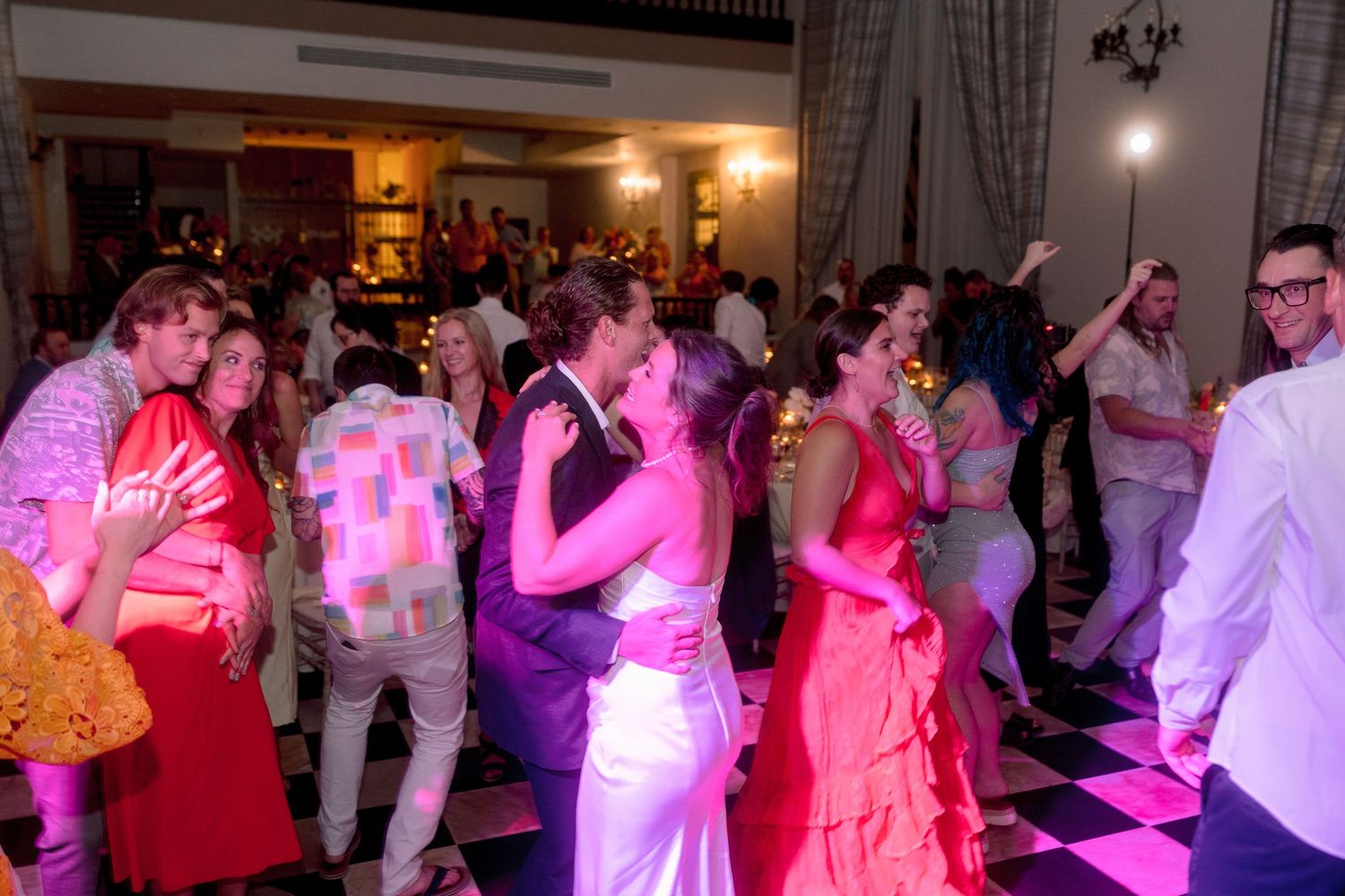 Couples dancing at a lively wedding reception with colorful lighting on a checkered dance floor.