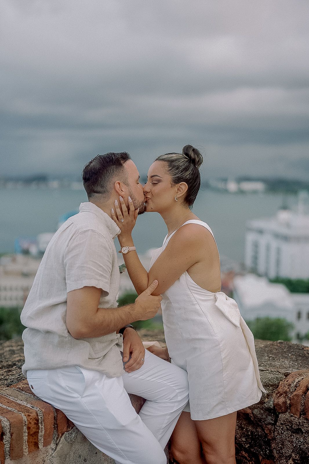 Couple sharing a romantic kiss on a stone wall with ocean and cloudy sky backdrop, dressed in white outfits.