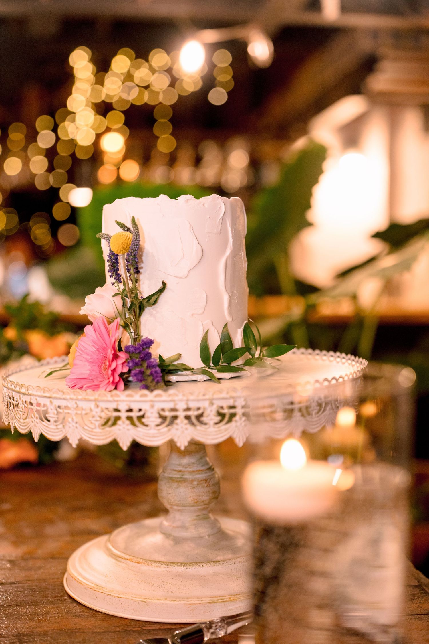 Small white wedding cake with pink and purple flowers on a vintage pedestal stand, with bokeh lights in background.