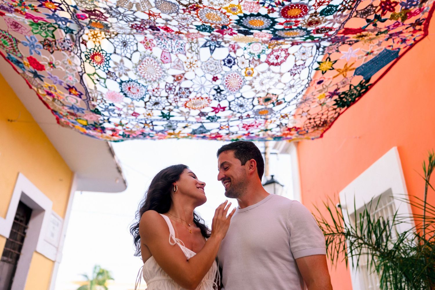 A couple smiles at each other under a colorful floral textile canopy on a vibrant outdoor patio.